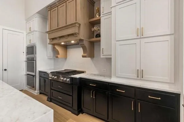 Gourmet kitchen featuring a stainless steel range, built-in double ovens, light wood-finish range hood, white and black cabinetry, and light-toned countertops