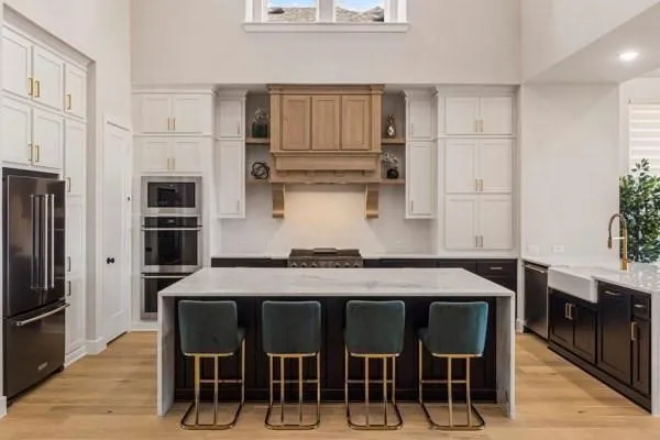 Two-tone cabinetry with gold-finish hardware, built-in stainless steel appliances, a large center island with a light-toned countertop, and wood-finish flooring