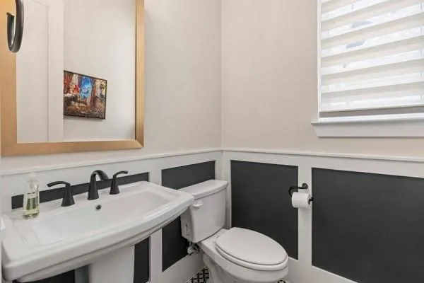 Pedestal sink with a matte black faucet, wall-mounted mirror with a gold-tone frame, two-tone wainscoting, and a window with blinds