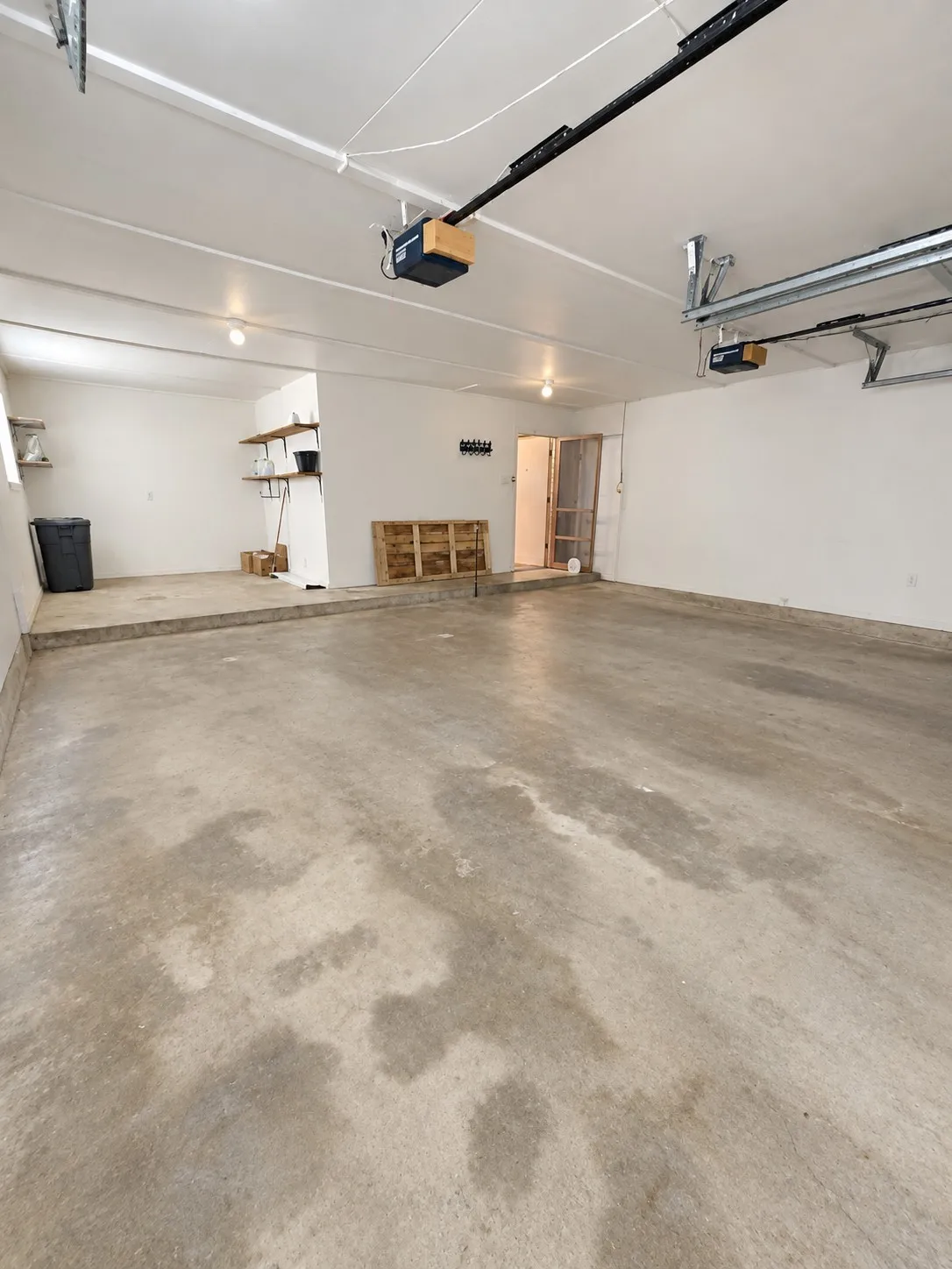 Concrete flooring garage with an elevated platform, exposed ceiling beams, built-in shelving, and garage door mechanisms
