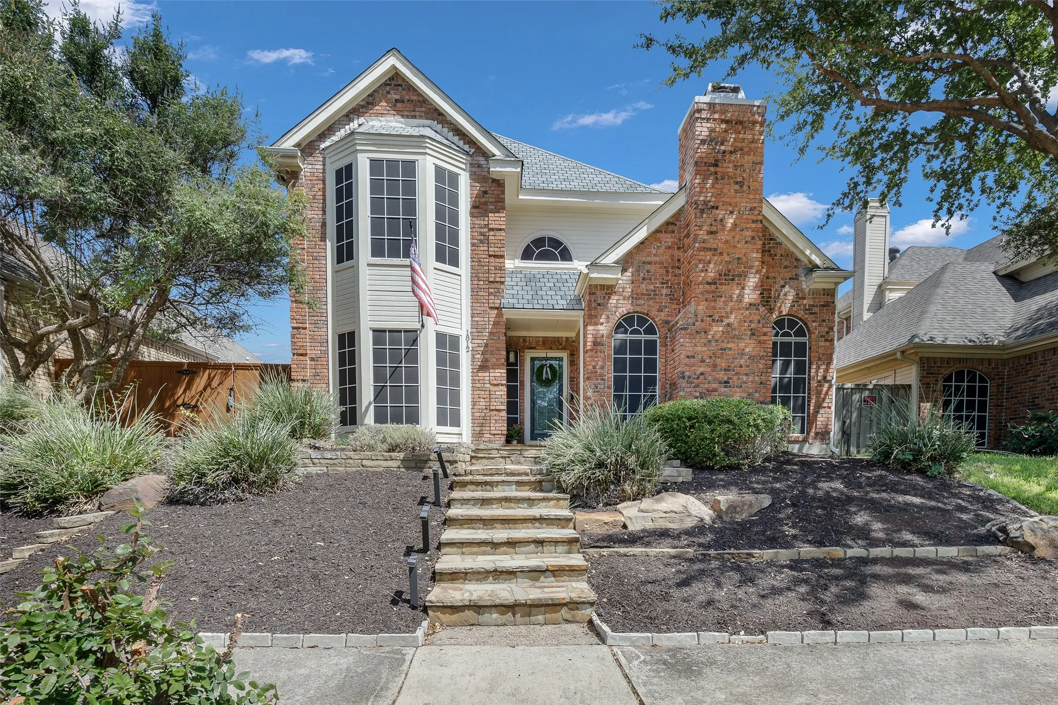 Brick facade with architectural bay window, stone-clad entry steps, integrated landscape lighting, and a prominent brick chimney