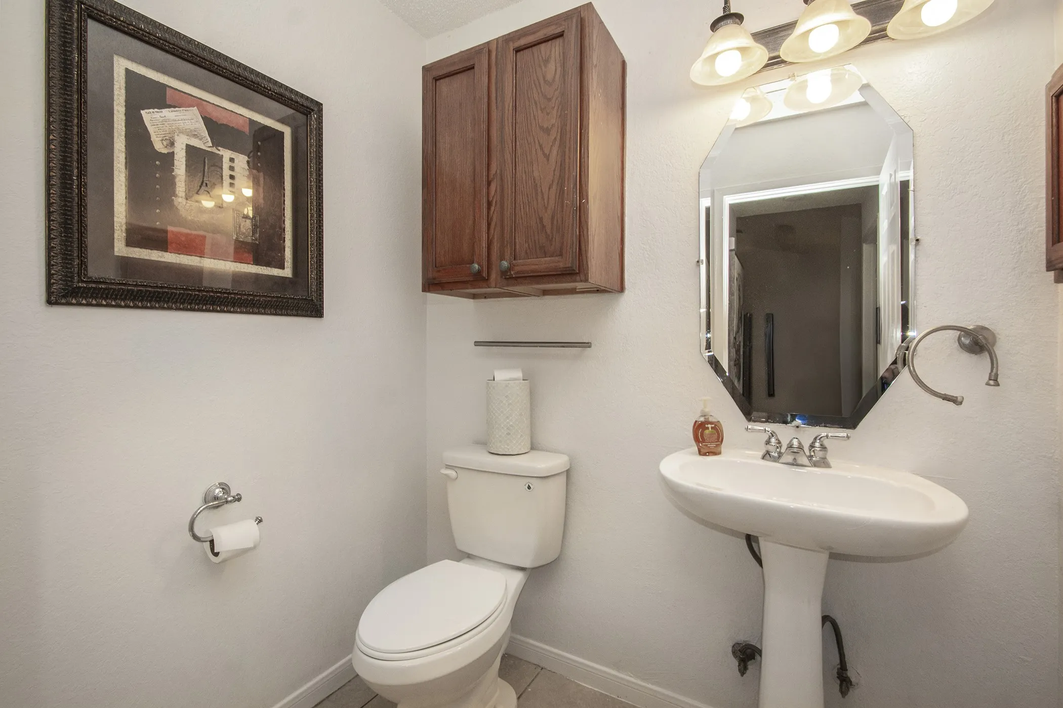 Pedestal sink with chrome fixtures, beveled octagonal mirror, three-light vanity fixture, and wall-mounted wood-finish cabinetry