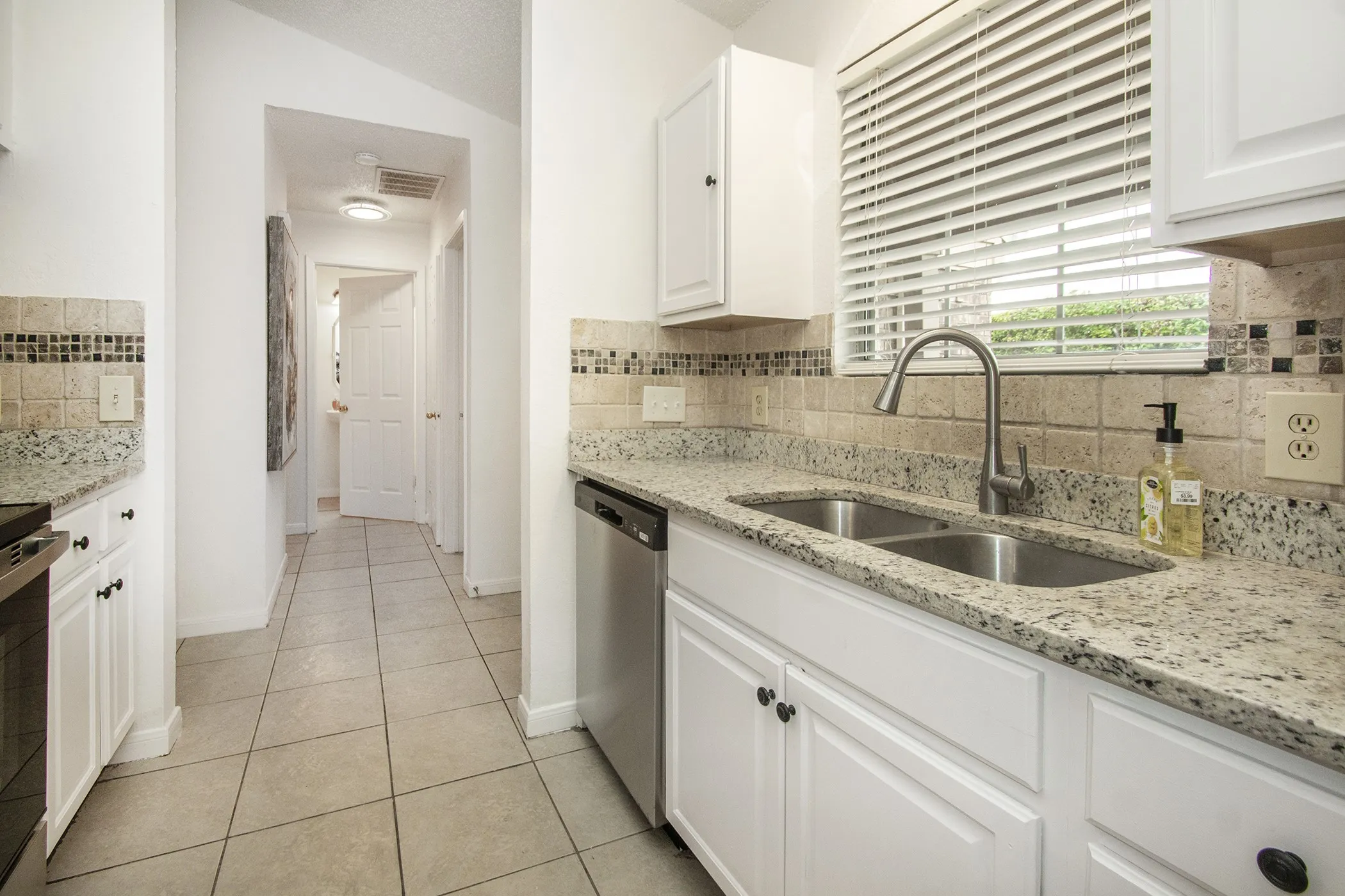 Kitchen featuring dual stainless steel sinks, a gooseneck faucet, light-toned countertops, white cabinetry with dark hardware, and a tiled backsplash