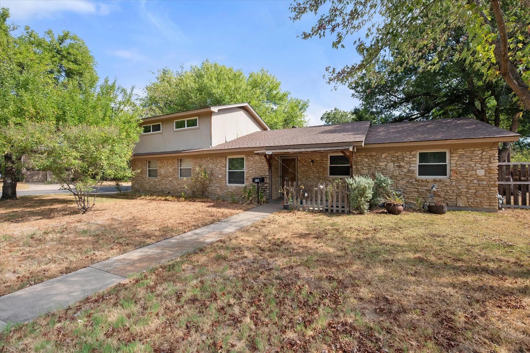 Stone veneer exterior with a concrete walkway, mature trees, and a partial picket fence