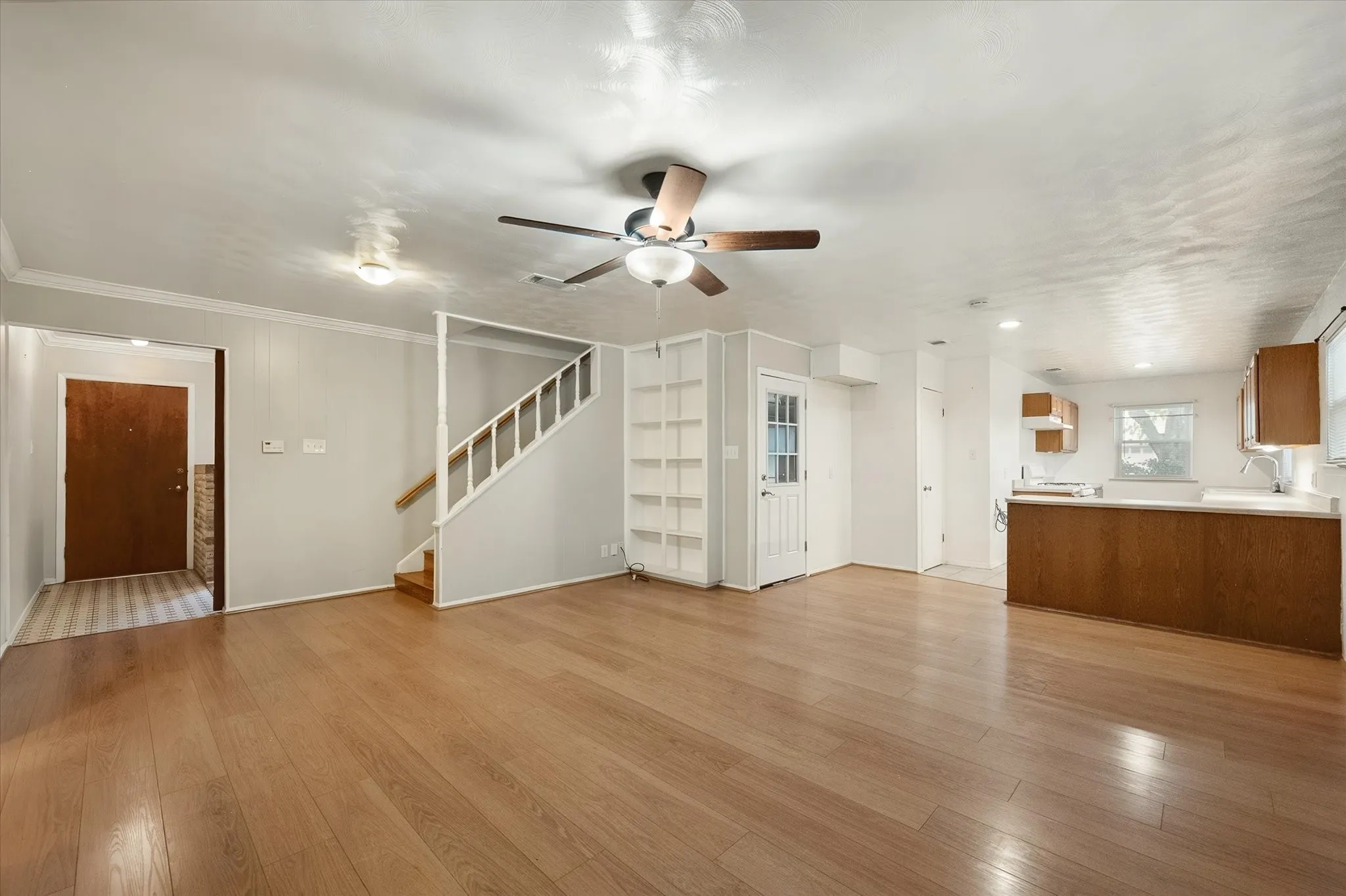 Wood-finish flooring, ceiling fan with integrated lighting, built-in shelving, staircase with white risers, and a kitchen featuring wood cabinetry