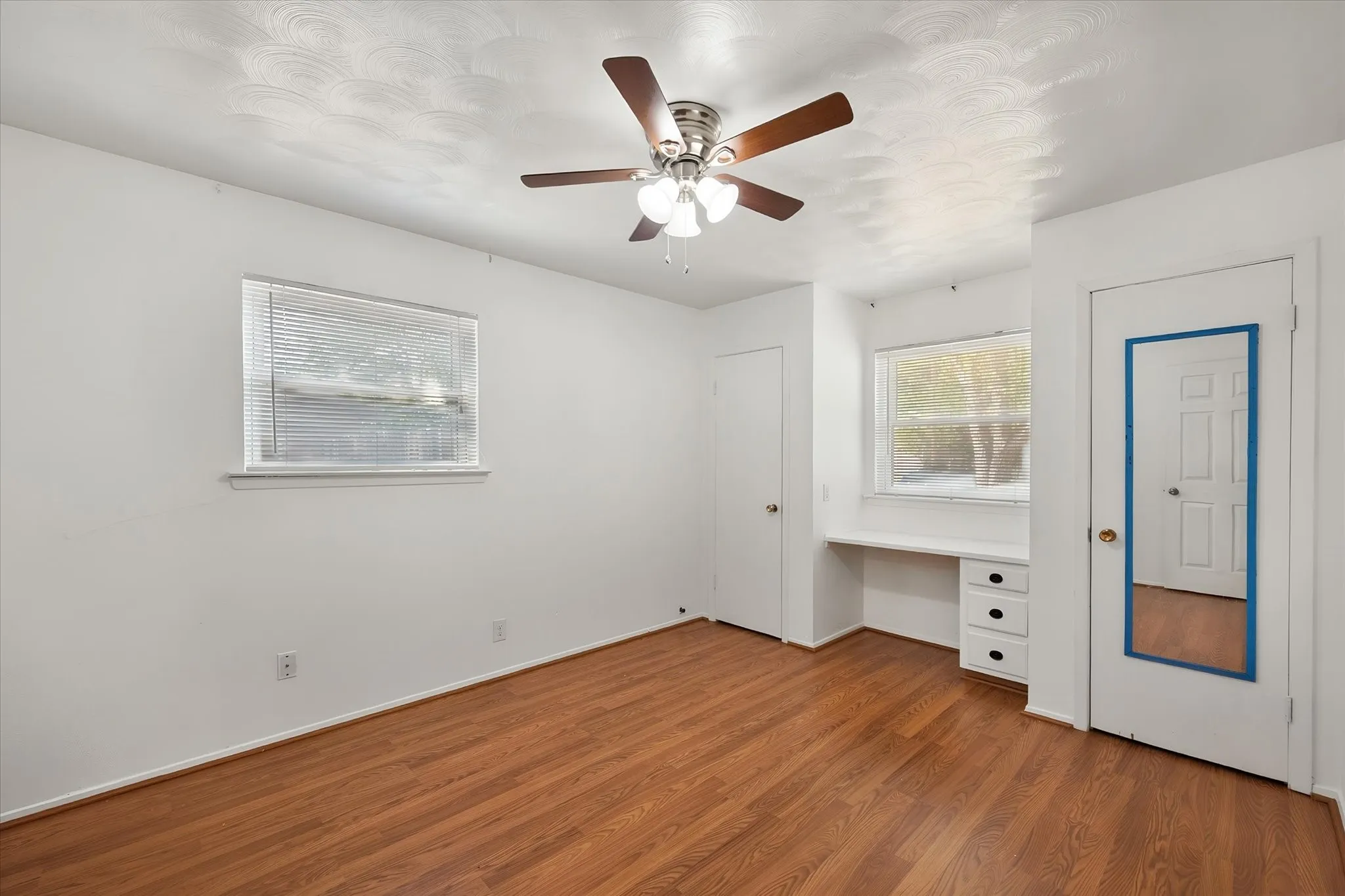 Wood-finish flooring, ceiling fan with integrated lighting, built-in desk with drawers, and two windows providing natural light