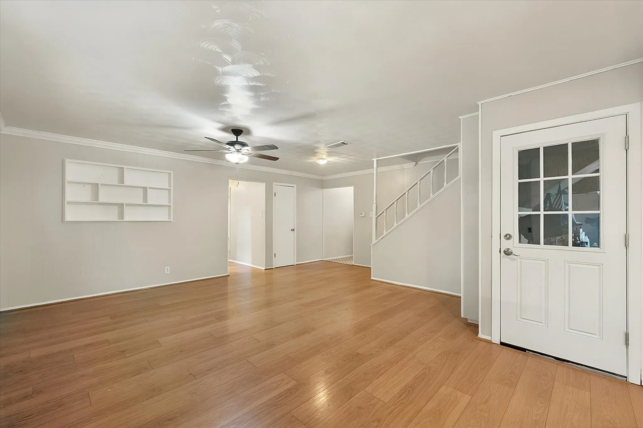 Wood-finish flooring, ceiling fan with integrated lighting, built-in wall shelving, crown molding, and a paneled door with glass inserts