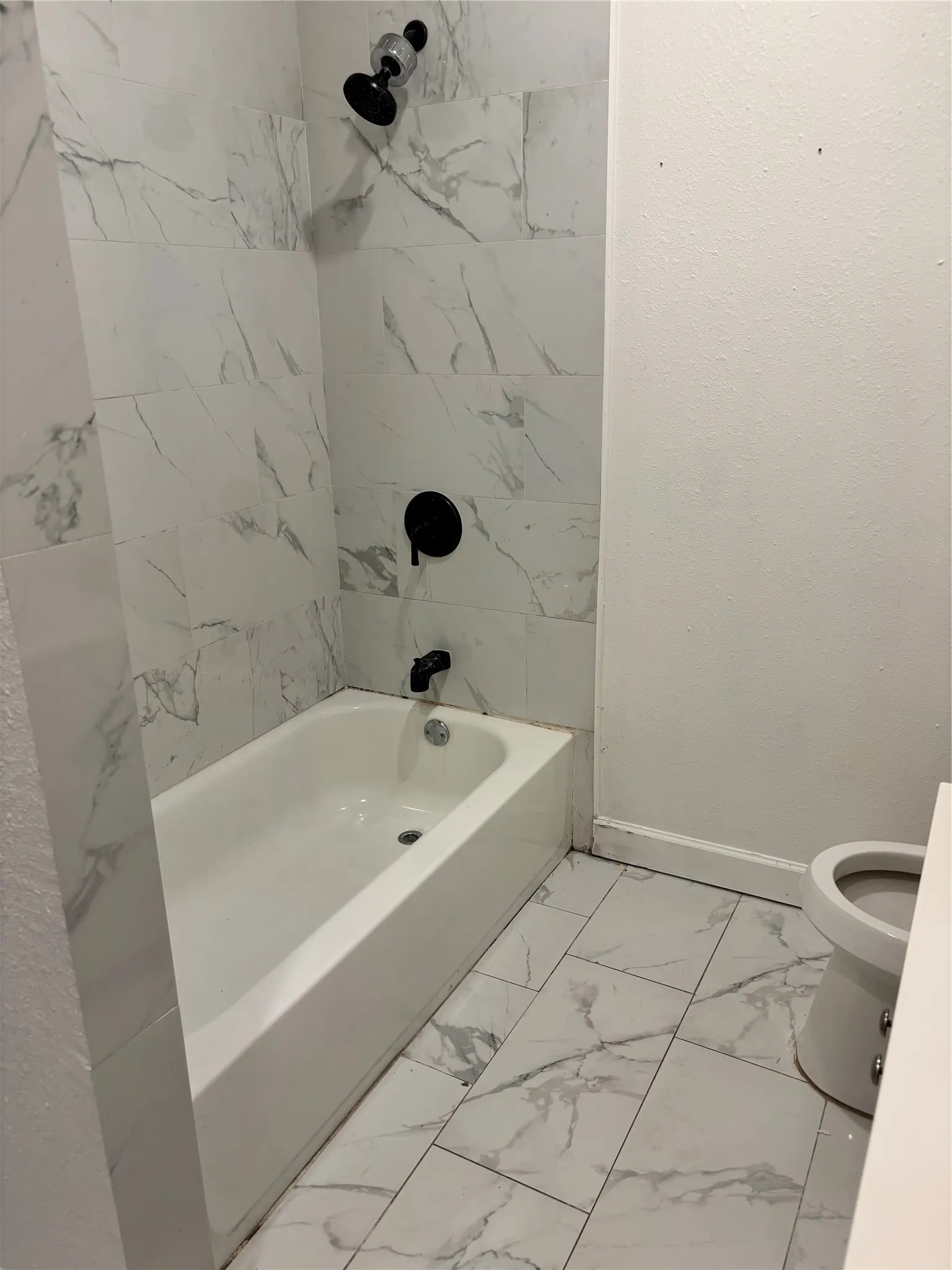 White subway tile shower surround with black matte fixtures, white bathtub, and coordinated tile flooring