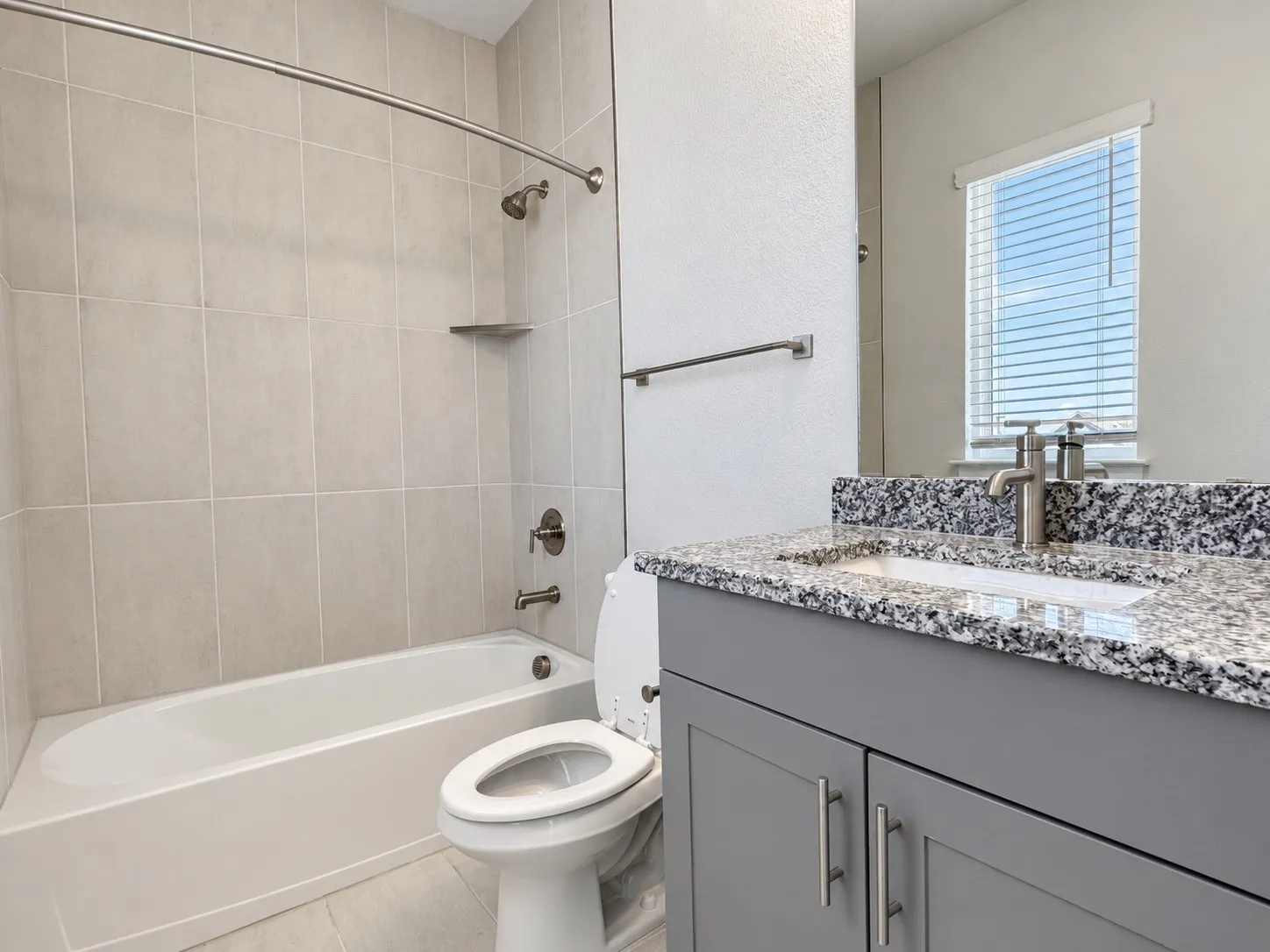 Bathroom featuring a white bathtub, tiled shower surround, gray vanity with granite countertop, and a window with blinds