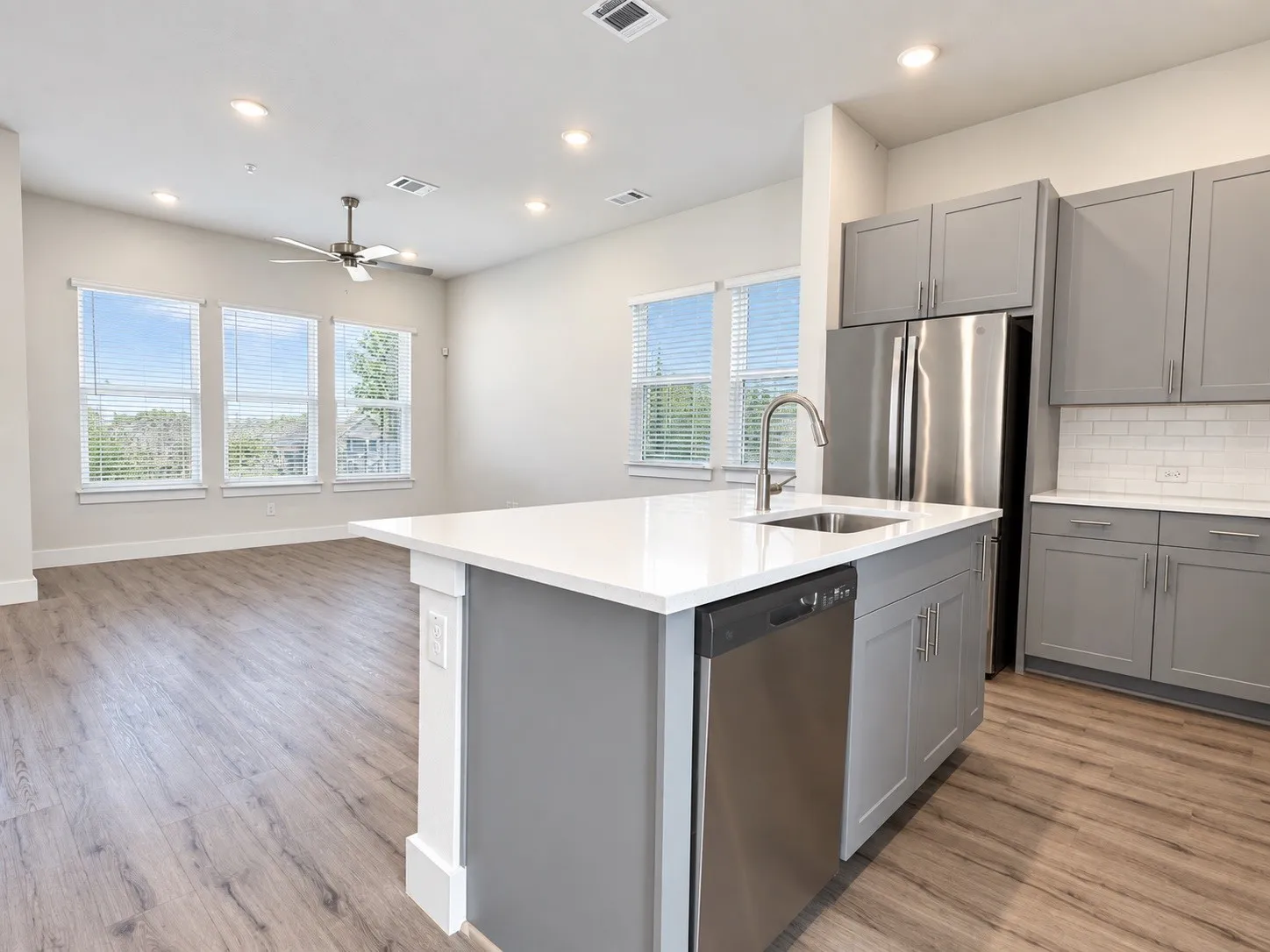 Kitchen island with a stainless steel sink and gooseneck faucet, light grey cabinetry, white subway tile backsplash, and wood-finish flooring