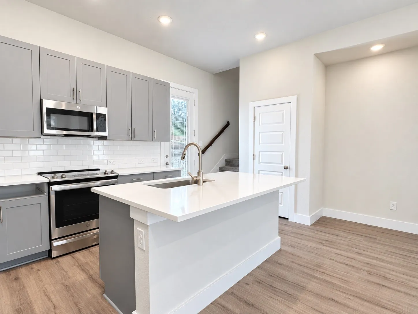 Kitchen featuring wood-finish flooring, gray cabinetry, white subway tile backsplash, stainless steel appliances, and a central island with a sink and white countertop