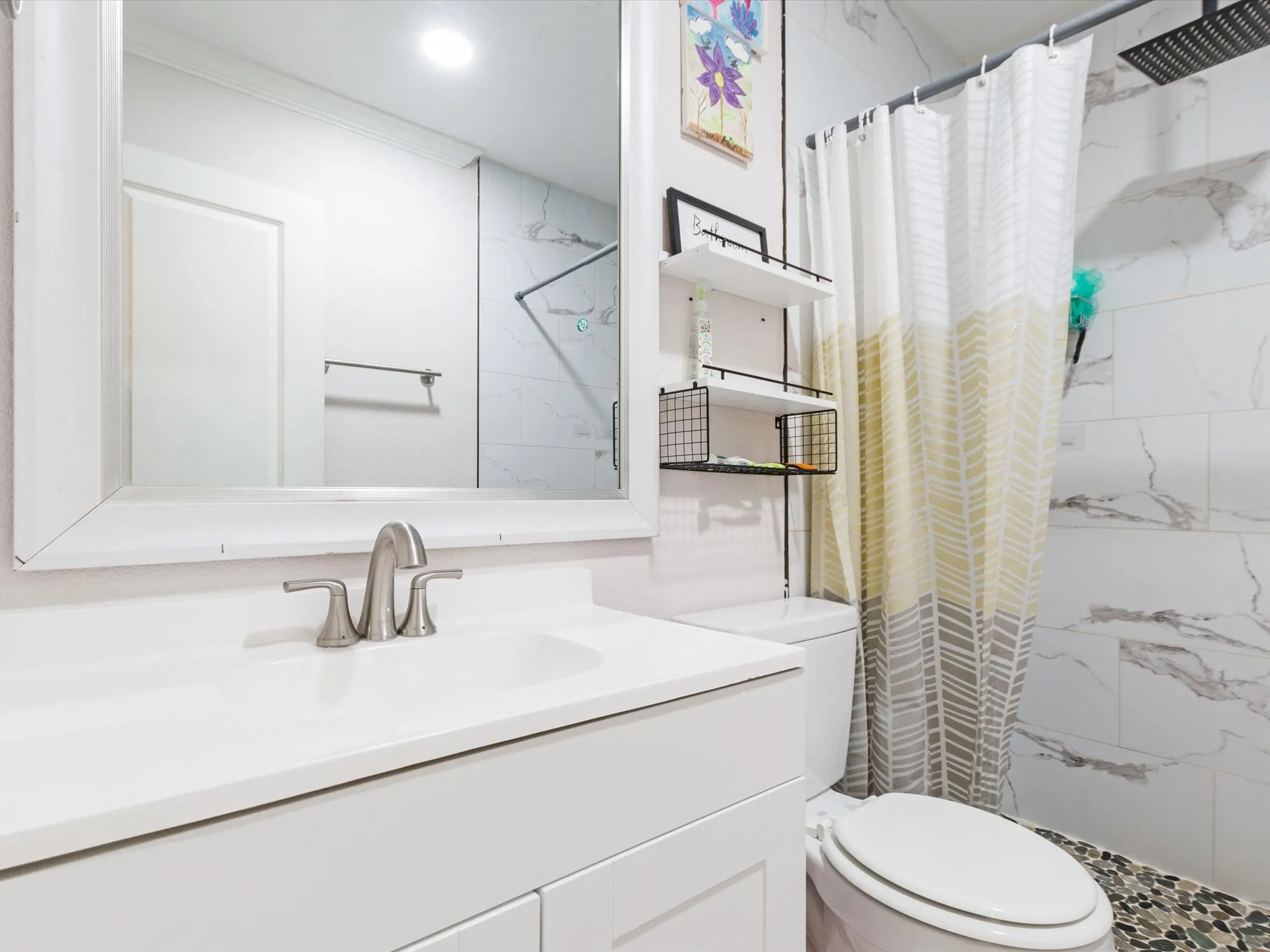White vanity with an integrated sink and brushed nickel faucet, large framed mirror, white toilet, and marble-look tile shower surround with a pebble-finish floor