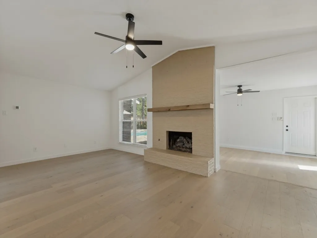 Vaulted ceiling with a ceiling fan, brick fireplace with a wood mantle, light wood-finish flooring, and a window with blinds