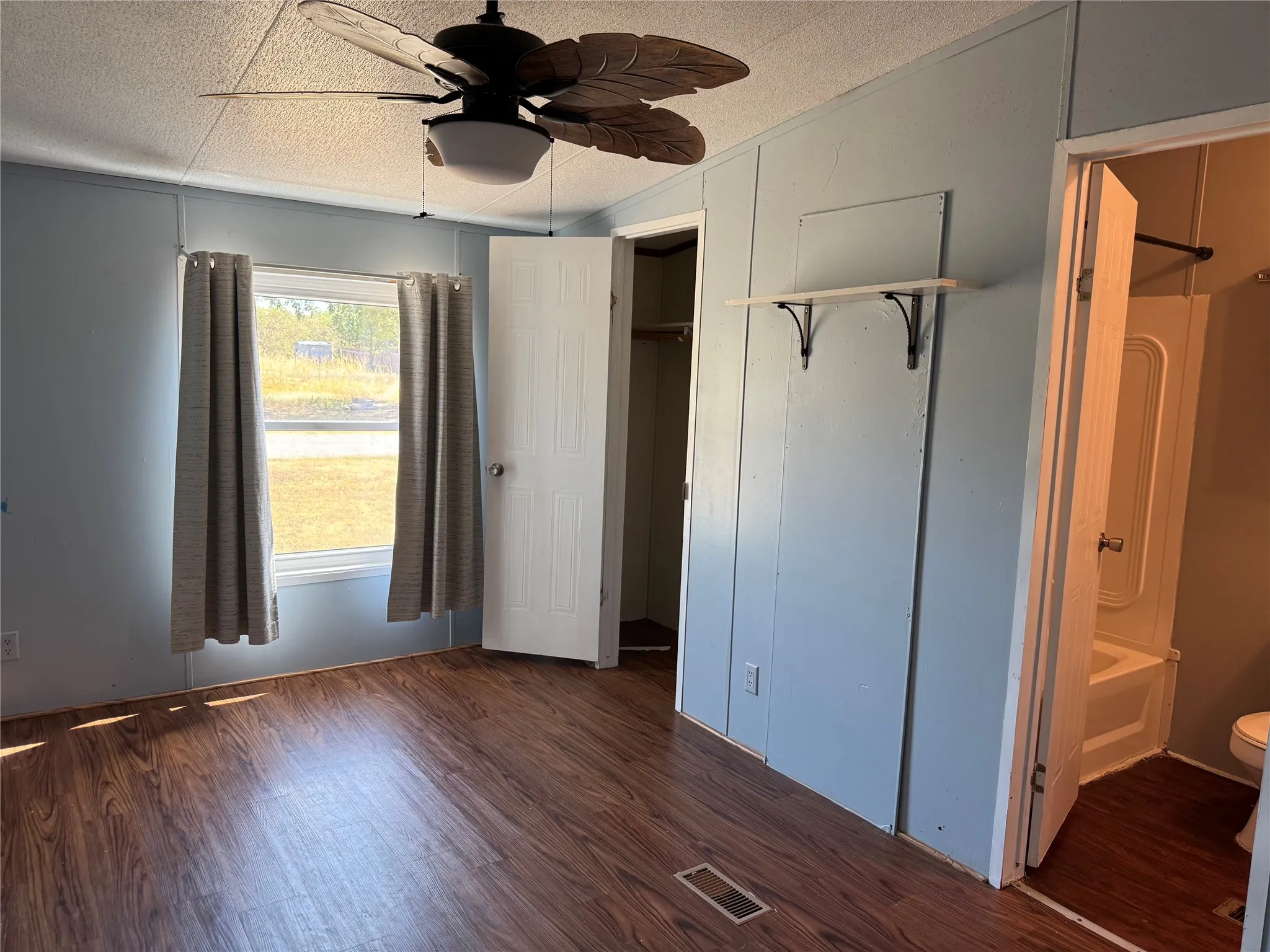 Wood-finish flooring, ceiling fan with integrated lighting, built-in shelving, white paneled doors, and a partial view of a bathroom with a white tub
