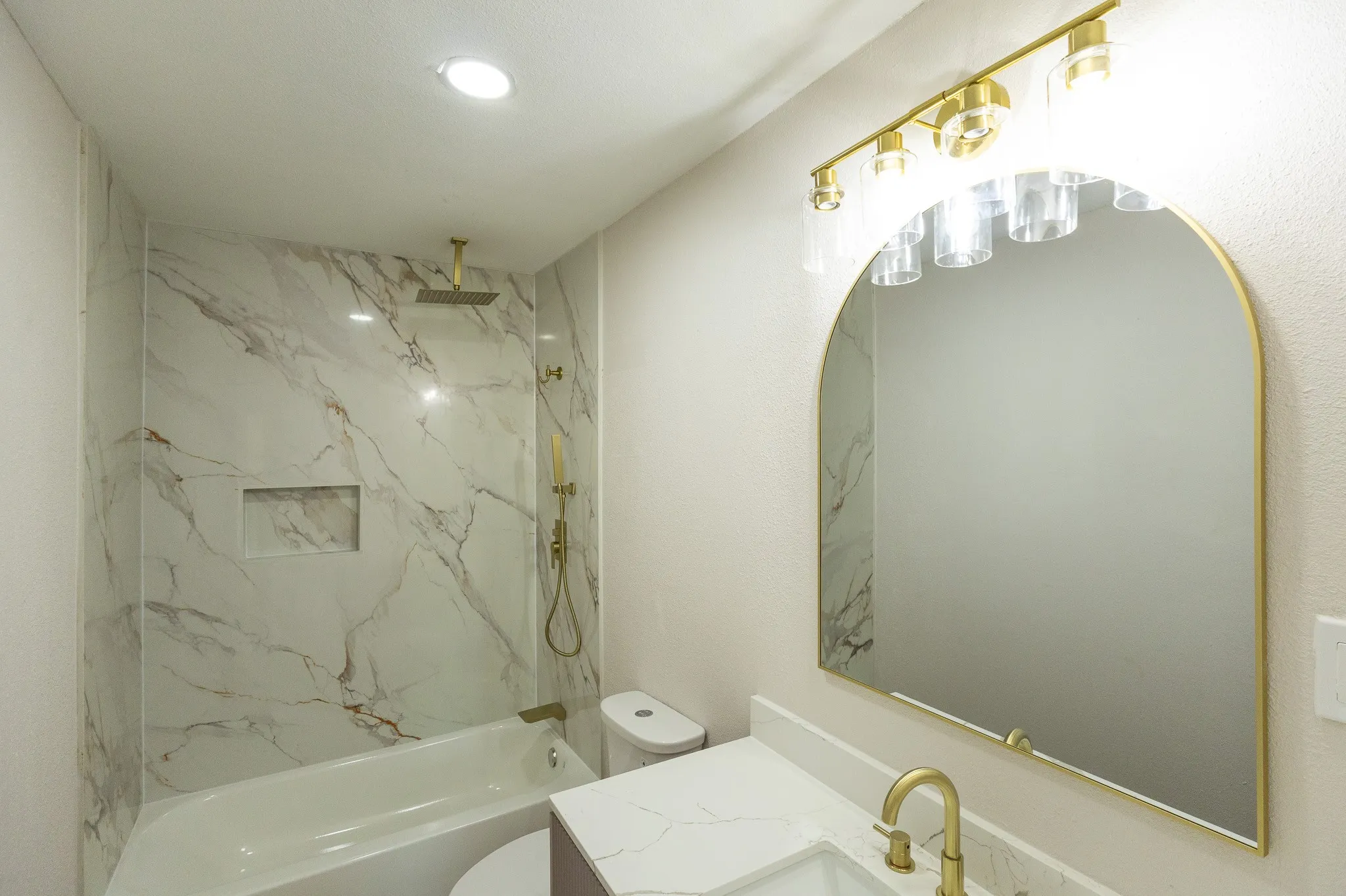 Bathroom featuring a white bathtub, gold-toned shower fixtures, a tiled shower surround with a built-in niche, a vanity with a white countertop, and a gold-framed arched mirror