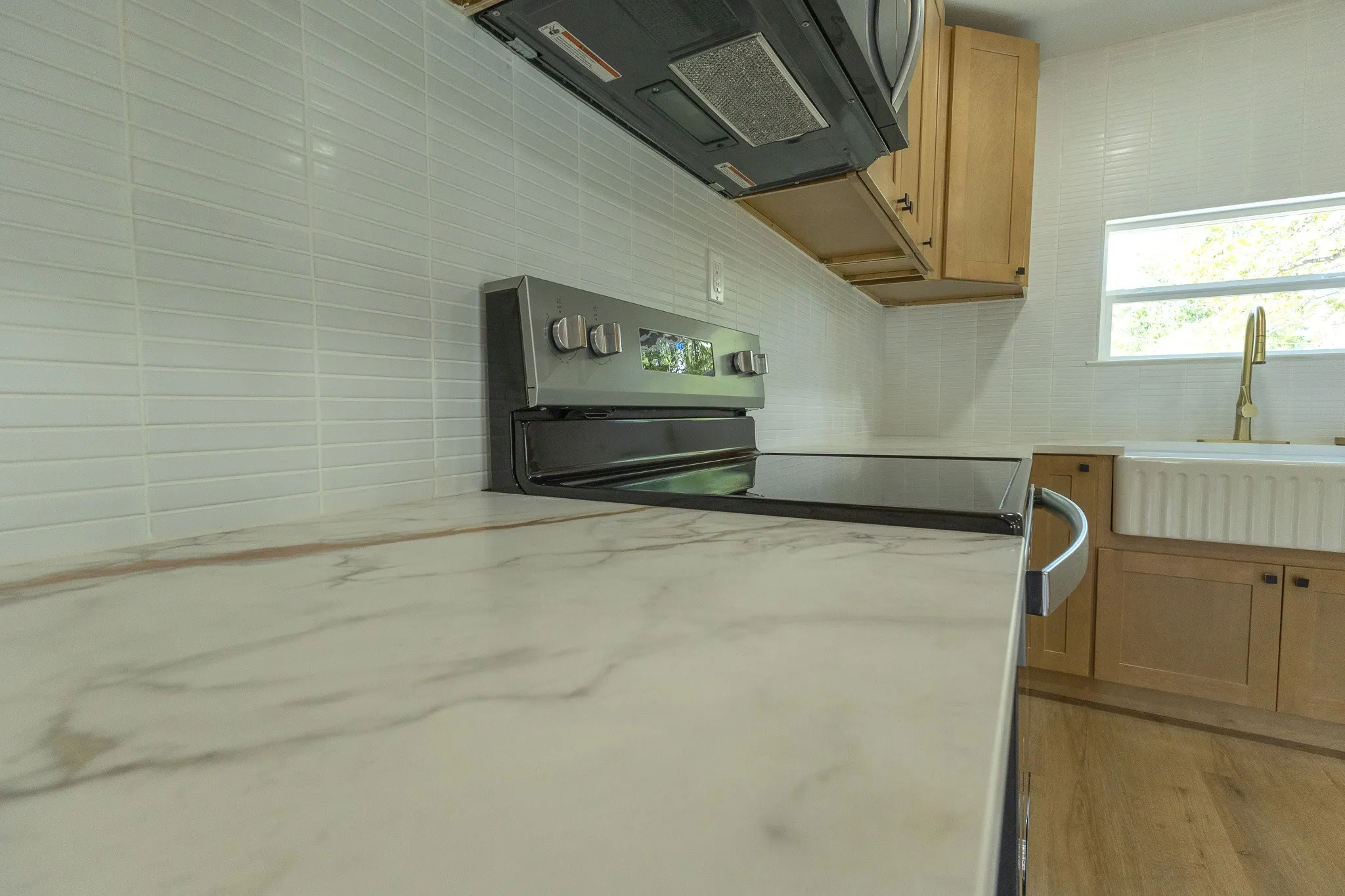 Integrated electric range, light-toned wood-finish flooring, white tile backsplash, light-toned cabinetry, and a white farmhouse sink with a brass faucet