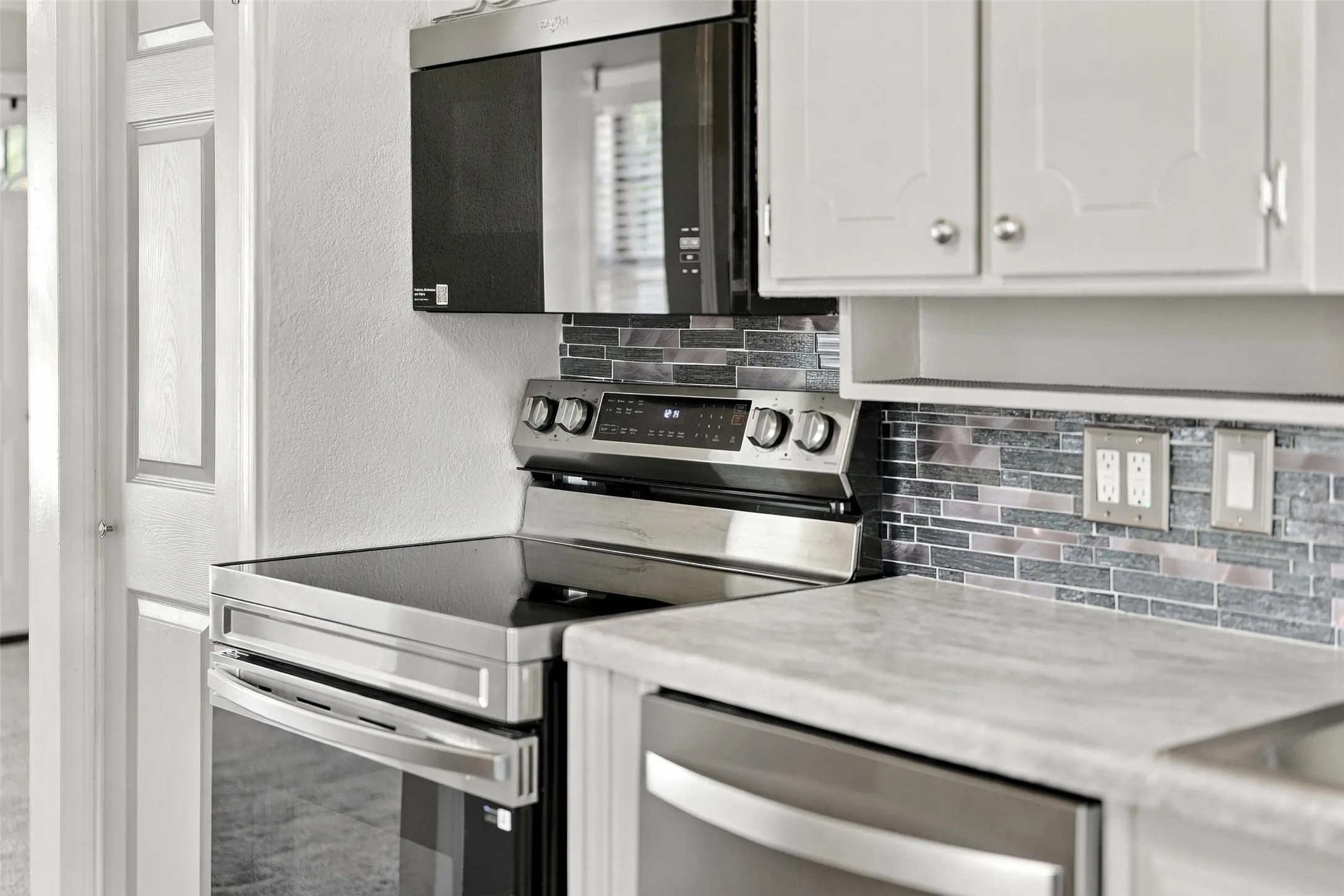 Stainless steel oven with a smooth cooktop, coordinating microwave, mosaic tile backsplash, light-toned countertops, and white upper cabinetry