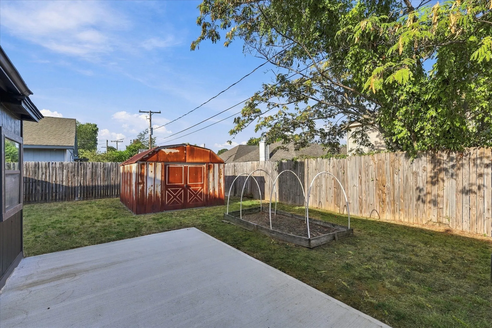 Spacious backyard oasis featuring a concrete patio, mature landscaping with established trees, wooden privacy fencing, a red storage shed with X-pattern detailing, and metal garden arbor structures ideal for climbing plants
