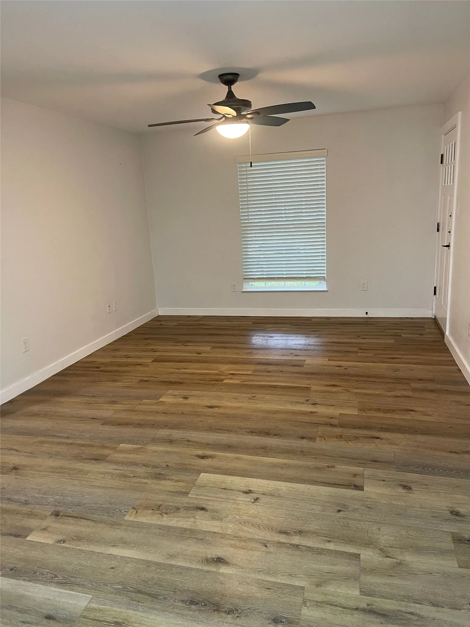 Spacious room featuring wood-finish flooring, a ceiling fan with integrated lighting, a window with blinds, and white walls