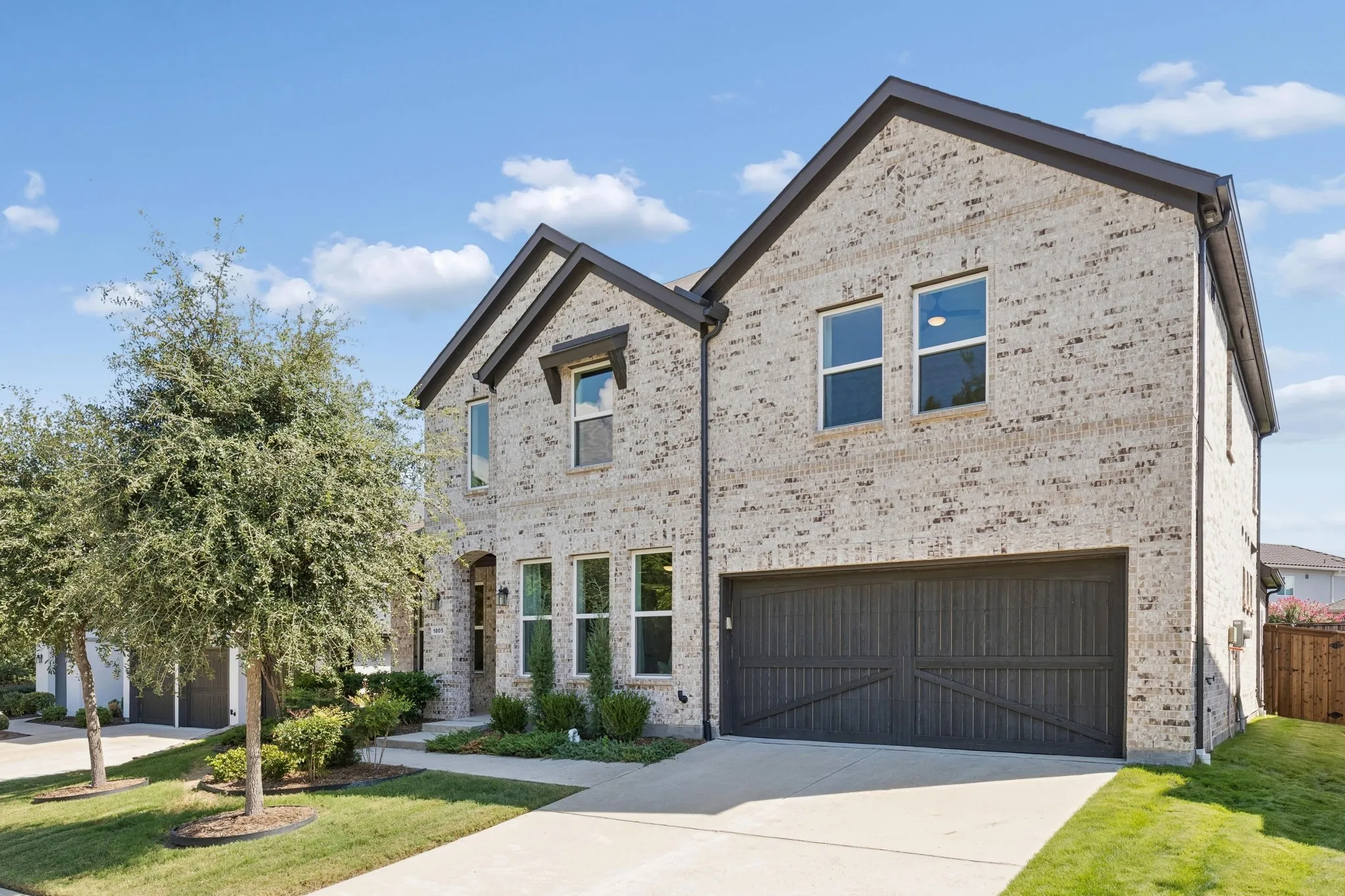 The handsome brick façade continues around the home with clean architectural lines, multiple gables, and a substantial wood-style garage door. Carefully landscaped beds and a spacious driveway provide both visual appeal and everyday functionality.