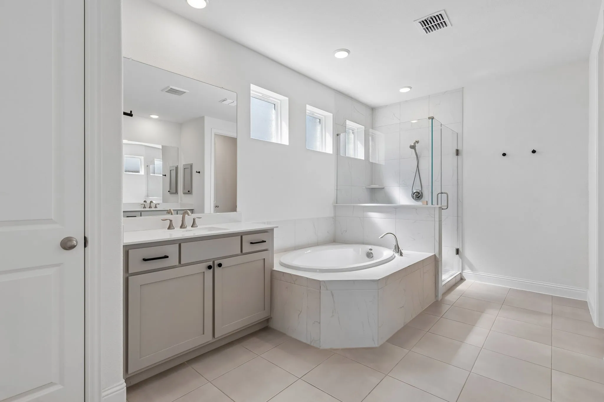 Bright and beautifully appointed, the primary bath offers a generous dual vanity, abundant cabinetry, a deep soaking tub, and a glass-enclosed shower accented by elegant marble-look tile. Multiple windows bring natural light into this spacious and refreshing retreat.