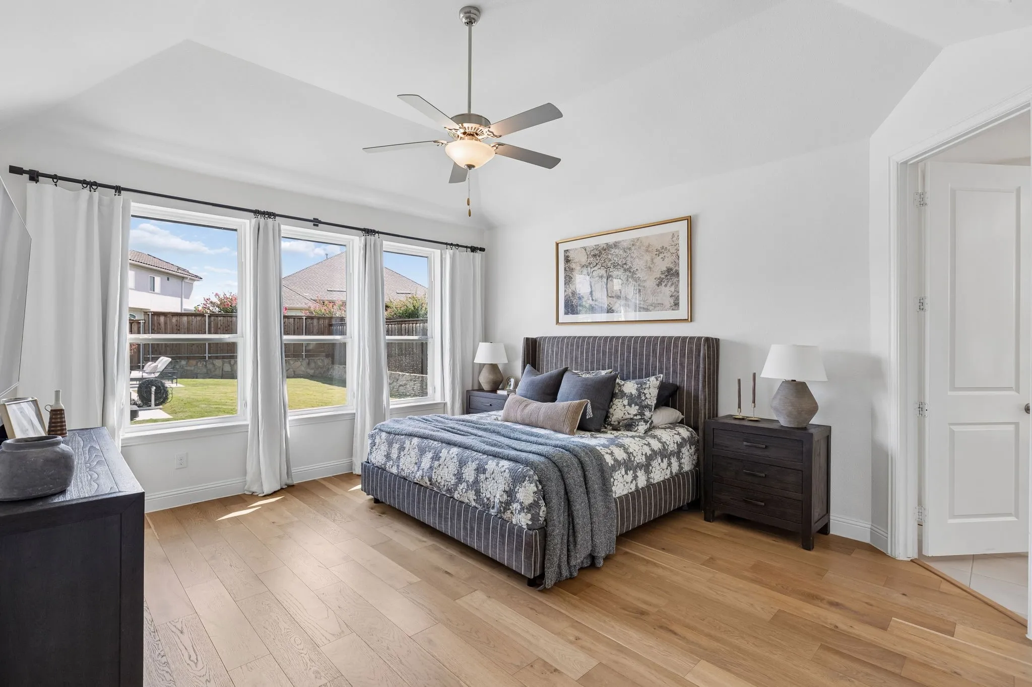 A serene retreat, the spacious primary bedroom is filled with natural light from a wall of windows overlooking the backyard. Warm wood floors, a tray-style ceiling, ceiling fan, and thoughtfully layered furnishings create a peaceful and inviting atmosphere.