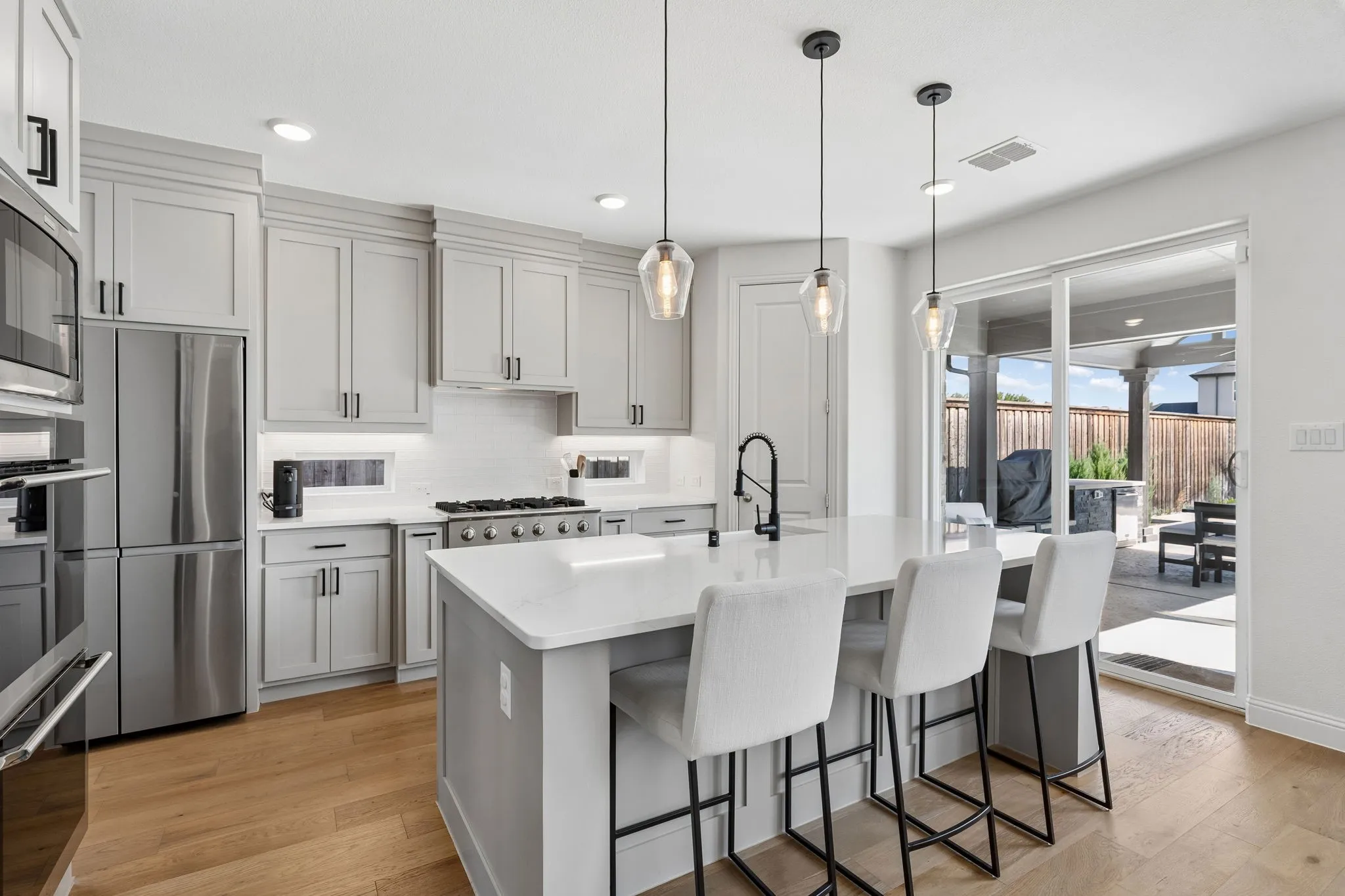 Sun-filled kitchen features extensive cabinetry, generous quartz counter space, a large island, statement pendant lighting, and a seamless connection to the covered outdoor living area.