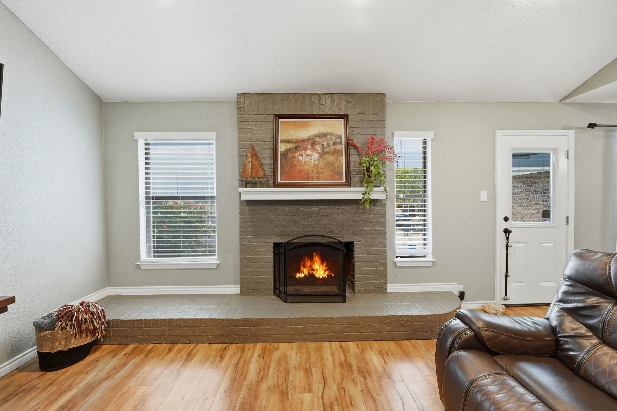 Wood-finish flooring with a brick fireplace, white mantel, and multiple windows with blinds
