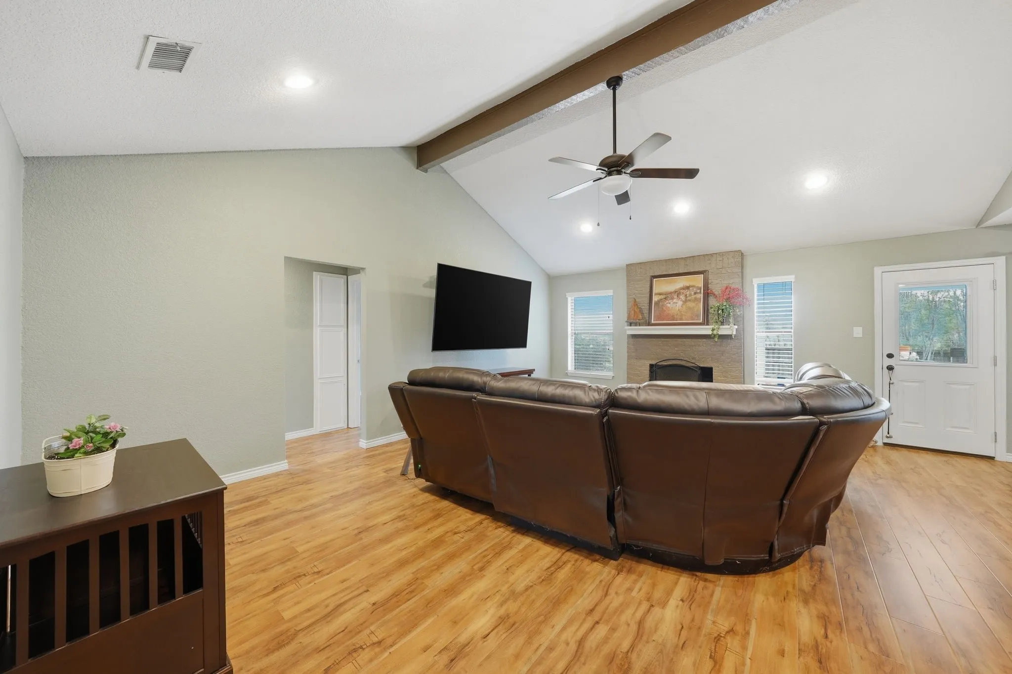 Vaulted ceiling with exposed beam, wood-finish flooring, brick-surround fireplace with mantel, recessed lighting, and ceiling fan