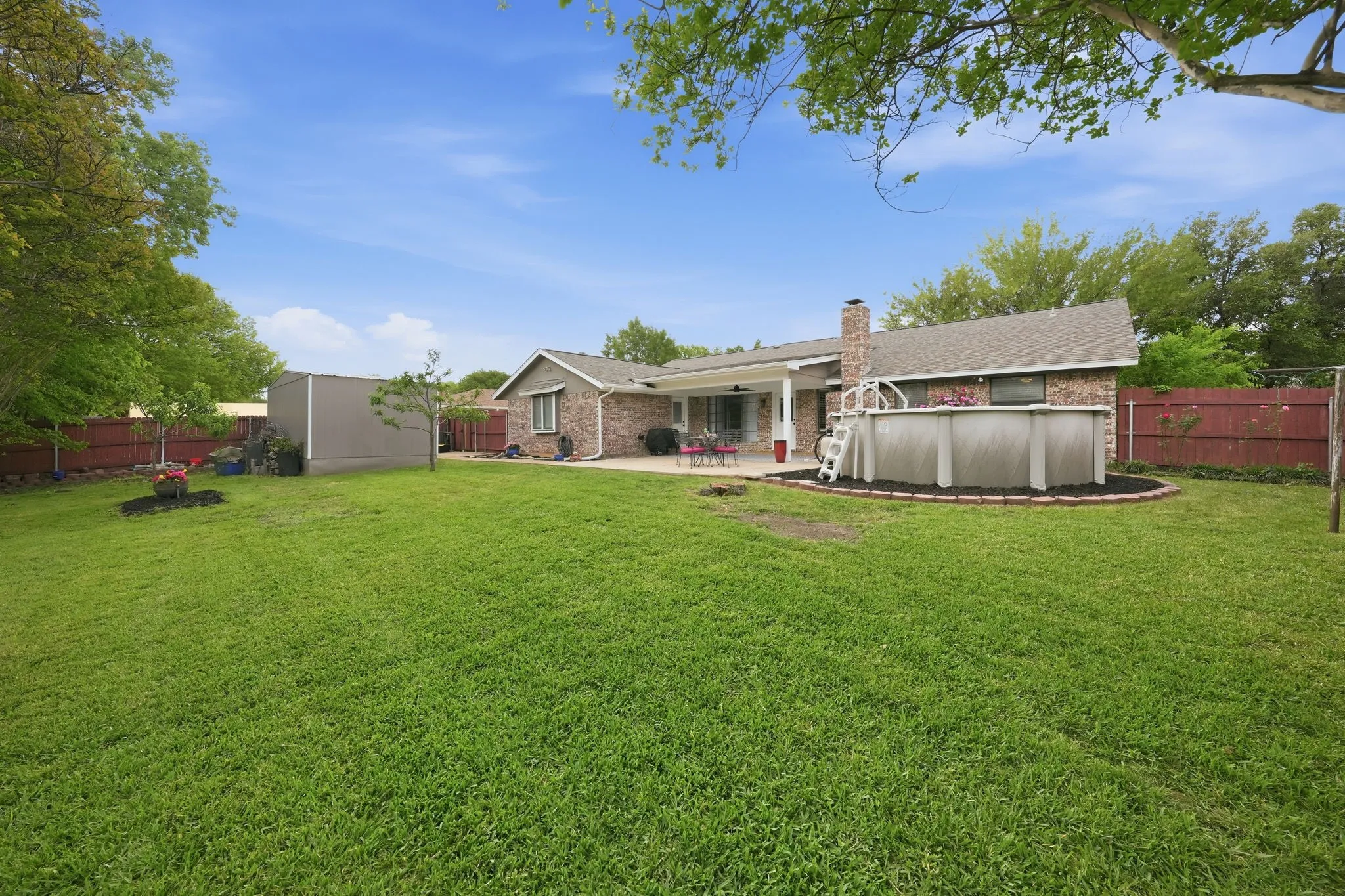 Expansive backyard featuring a manicured lawn, brick exterior, covered patio, above-ground pool, and a storage shed