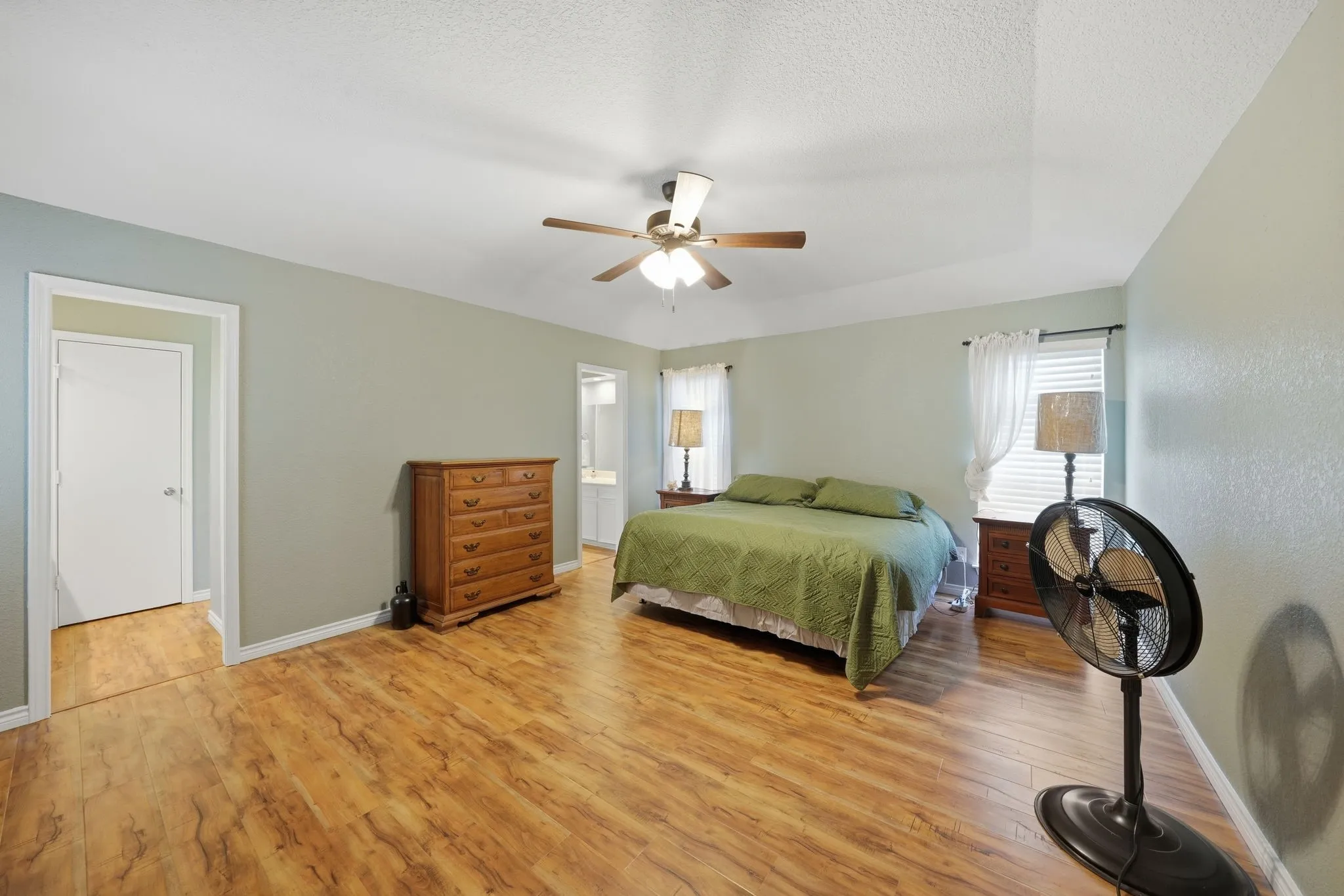 Spacious primary bedroom featuring wood-finish flooring, light wall paint, a ceiling fan, and white window blinds