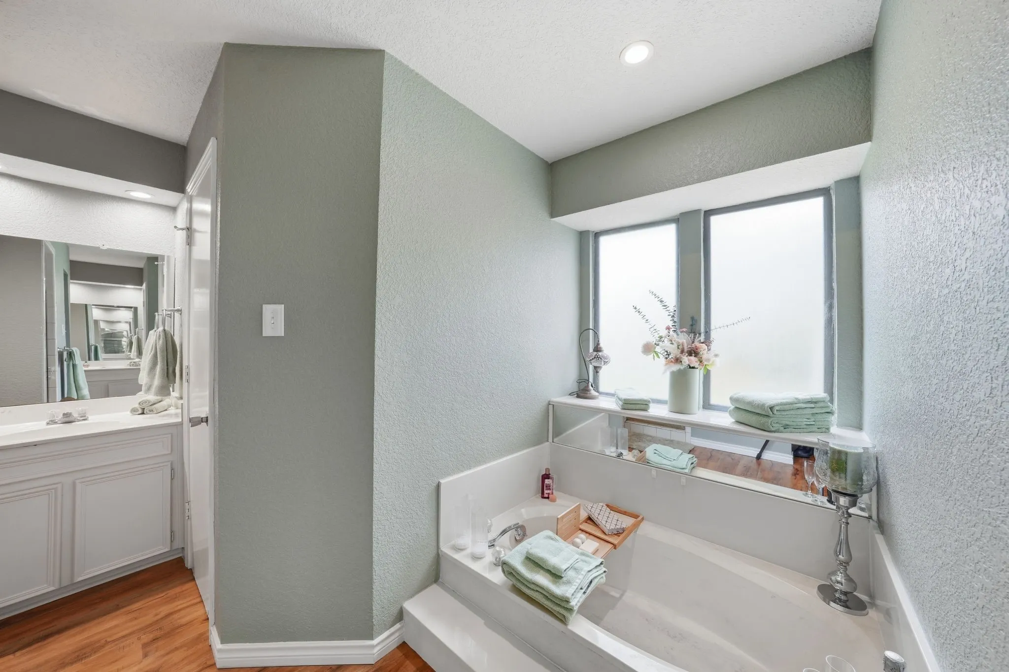 Built-in corner soaking tub with integrated shelving, frosted windows, recessed lighting, wood-finish flooring, and a vanity with a large mirror