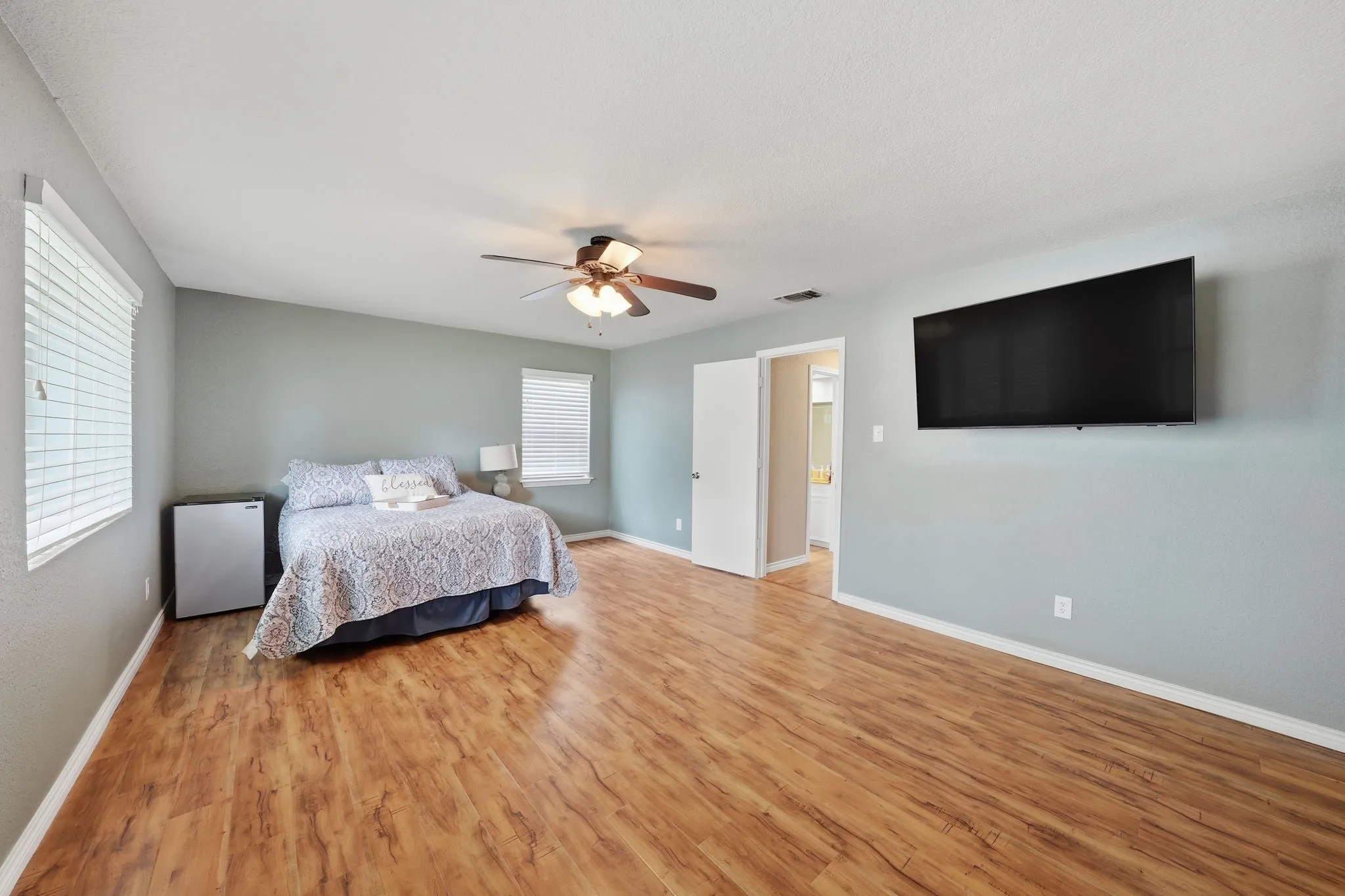 Spacious room featuring wood-finish flooring, light-toned walls, multiple windows with blinds, and a ceiling fan with integrated lighting