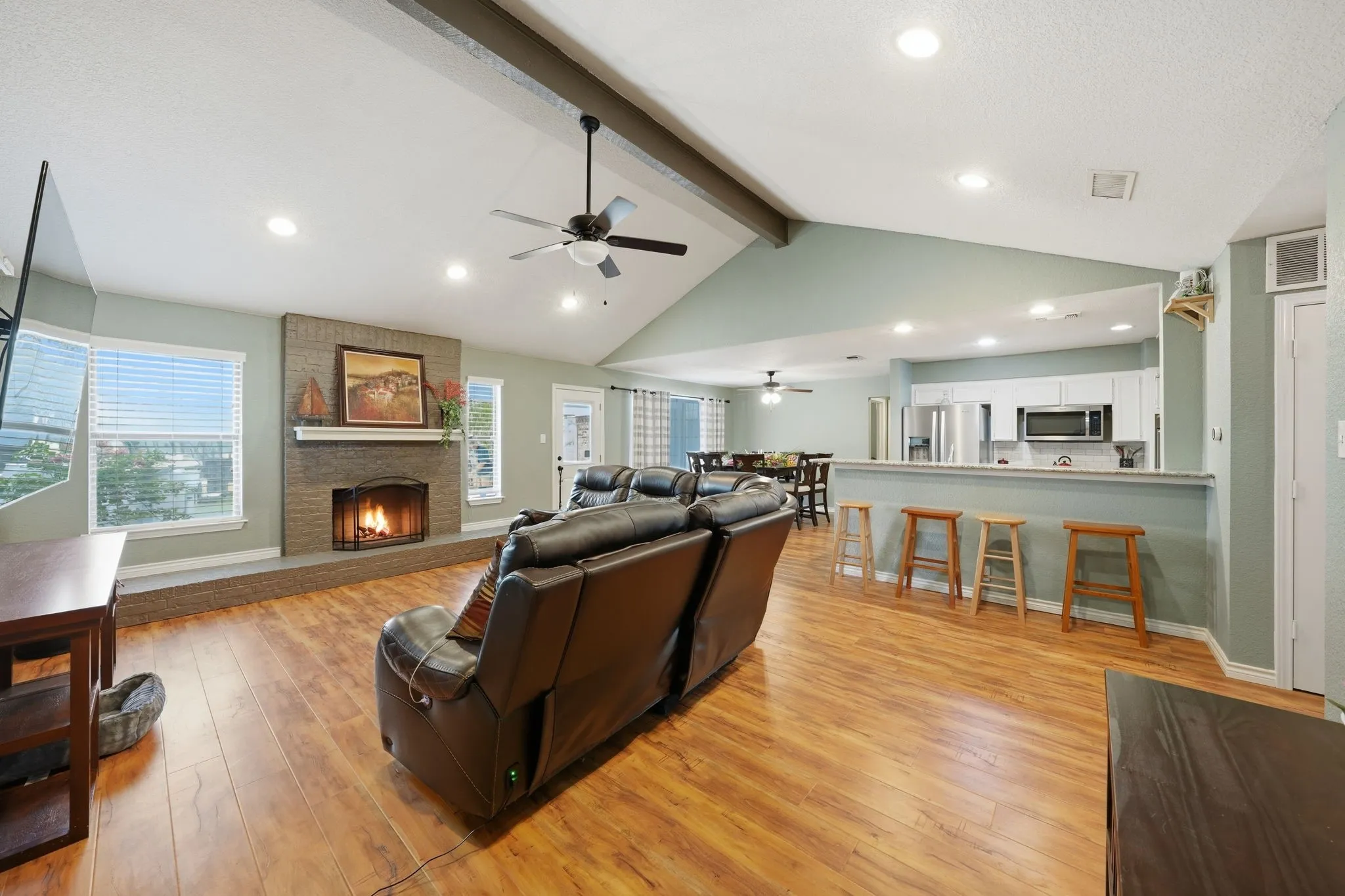 Vaulted ceiling with exposed beam, recessed lighting, wood-finish flooring, brick-faced fireplace, and a kitchen pass-through