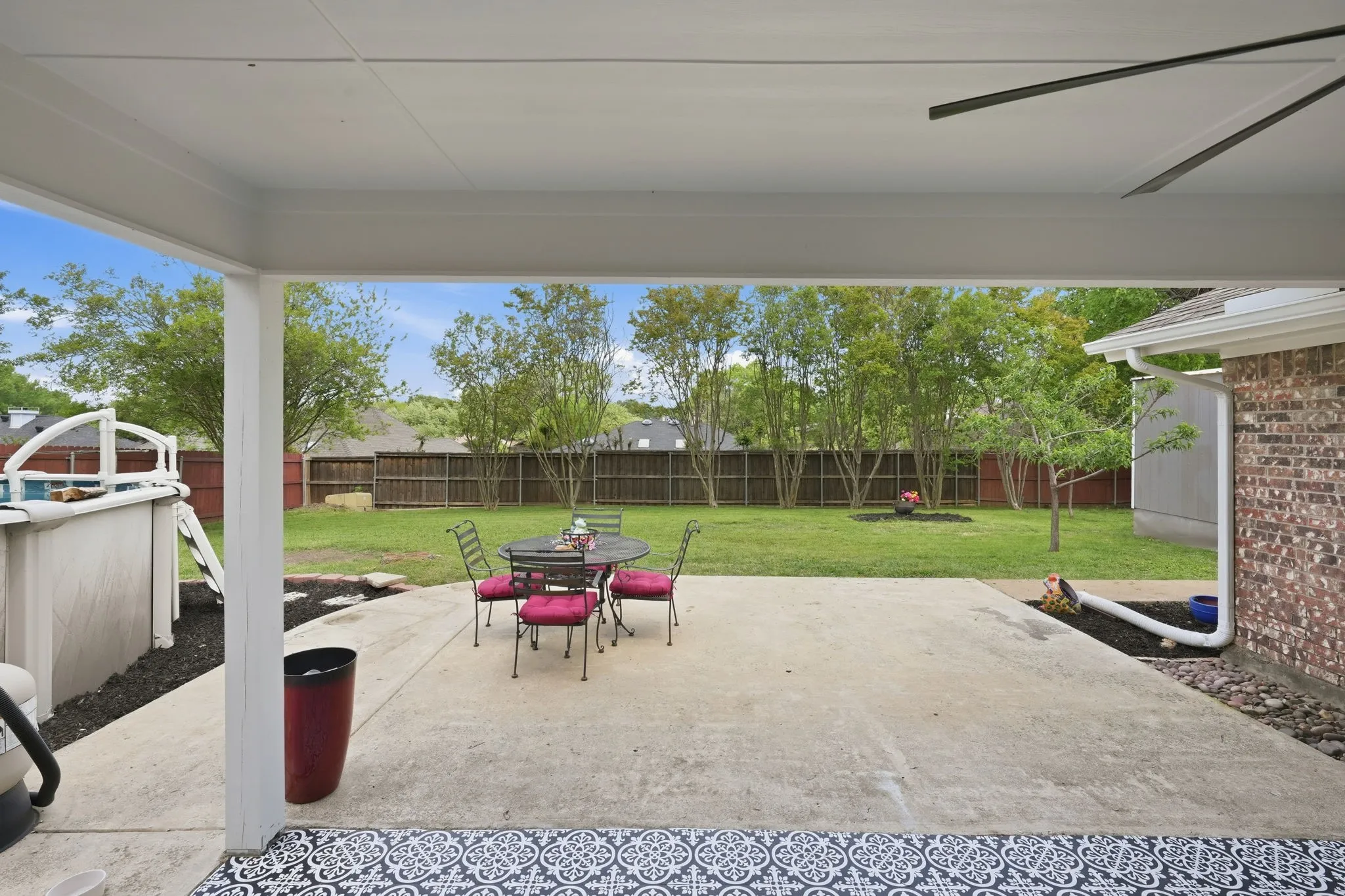 Covered patio with concrete flooring and decorative tile accents, offering an expansive backyard with a manicured lawn and mature trees