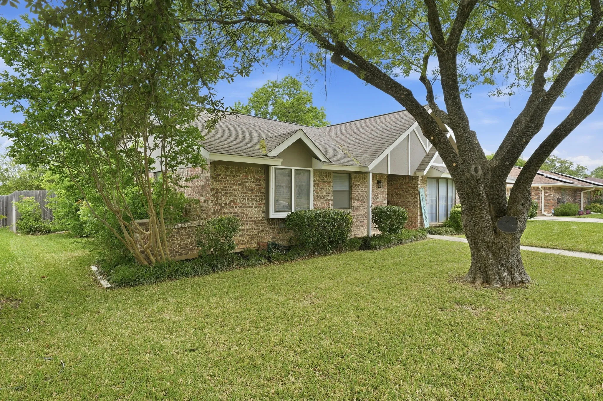 Brick exterior facade, gabled roofline, mature trees, landscaped shrubbery, and a side yard with a wooden fence