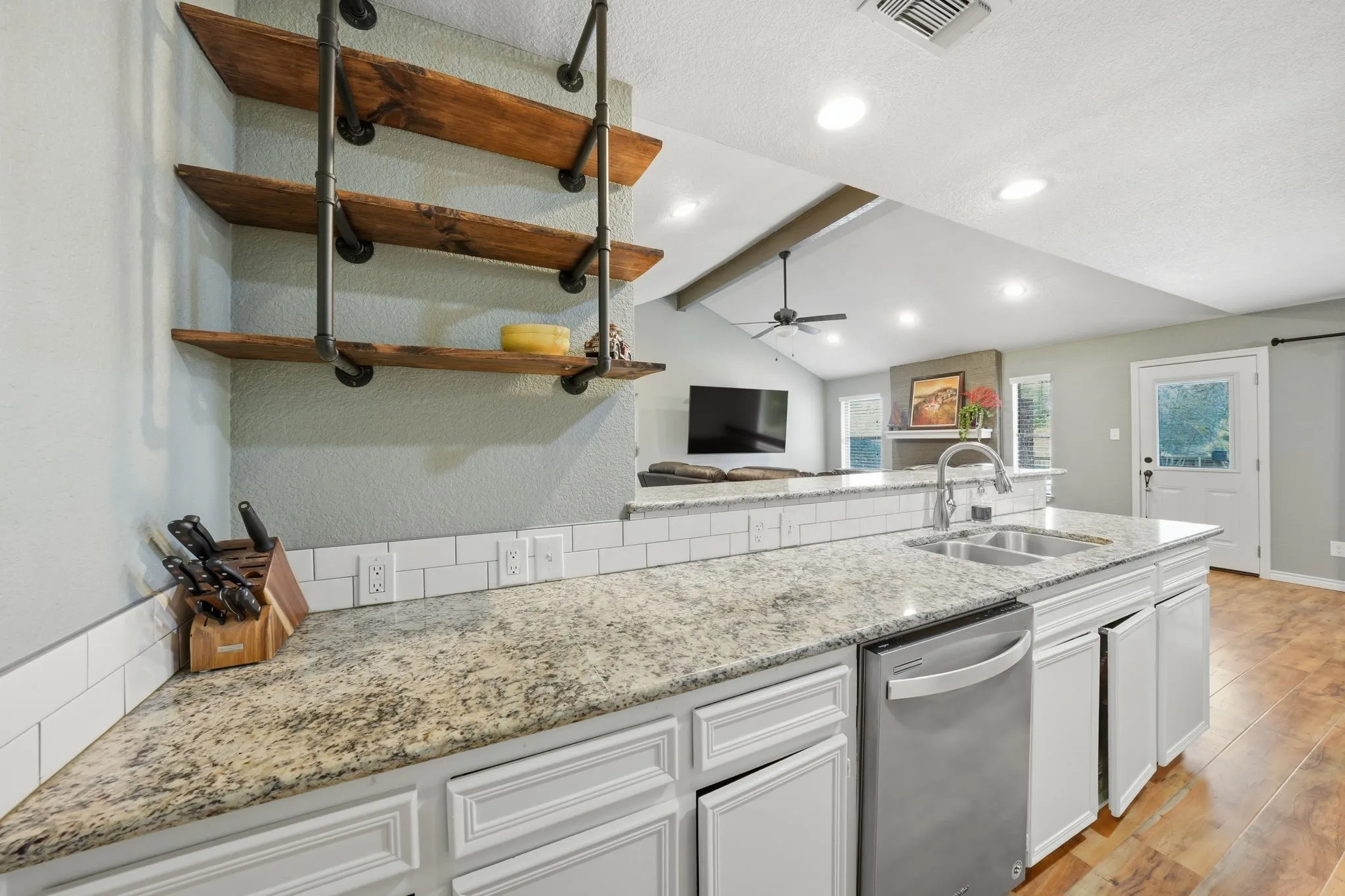 Kitchen featuring granite countertops, white subway tile backsplash, stainless steel dishwasher, and open shelving with wood and pipe accents