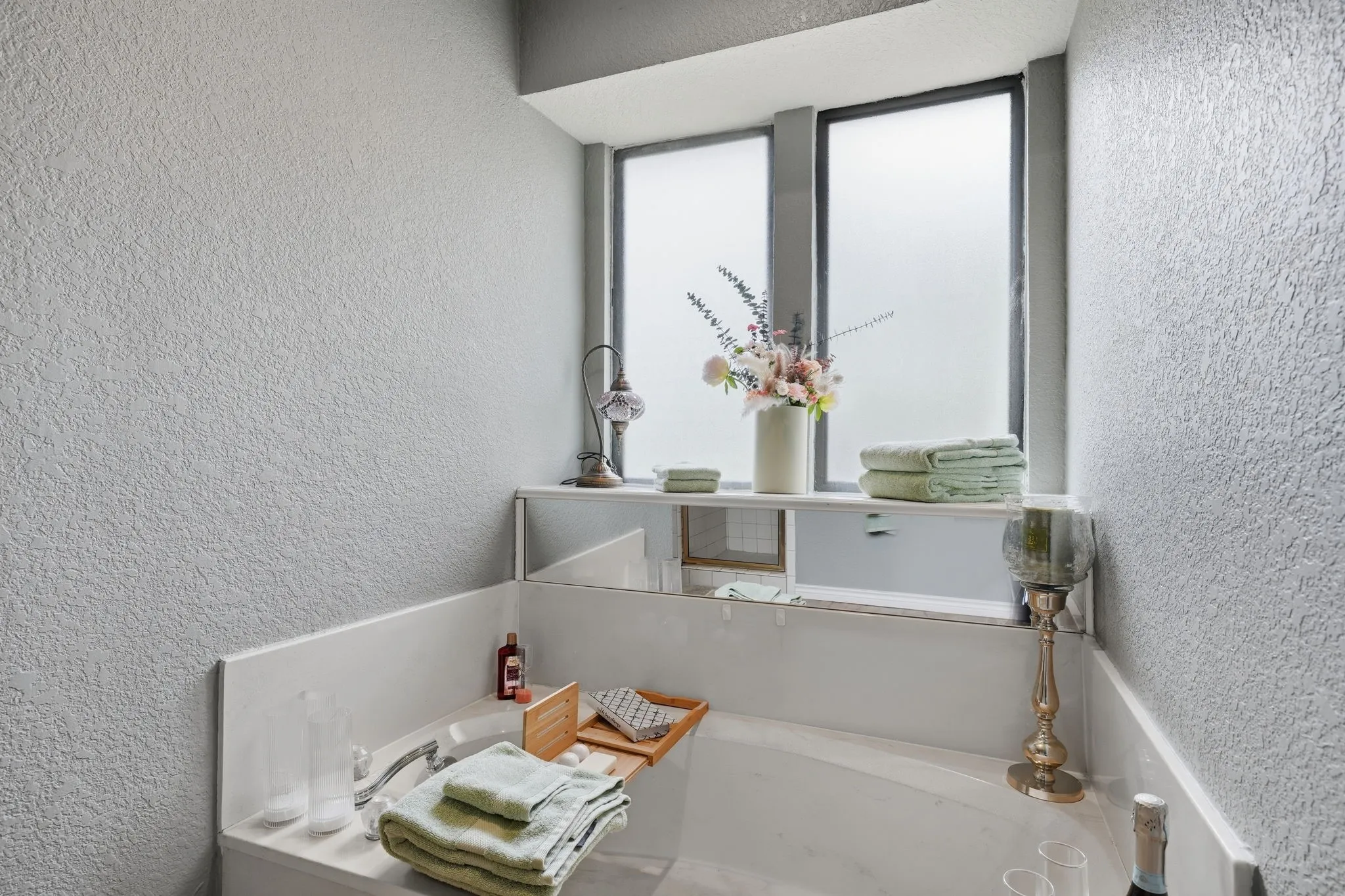 Recessed soaking tub with surrounding ledge, built-in shelving, textured wall finish, and frosted window panes