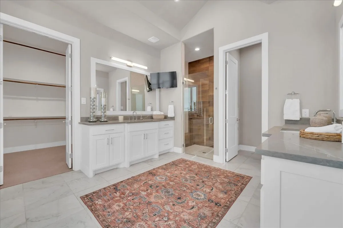 Bathroom with dual vanities featuring white cabinetry and grey countertops, a walk-in shower with glass enclosure and wood-finish wall, and a walk-in closet