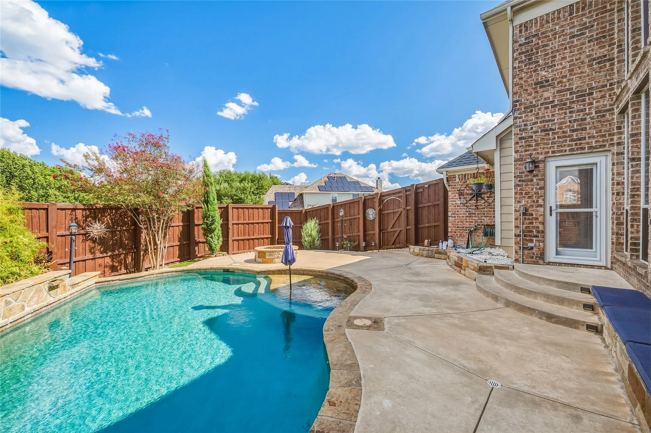 Outdoor living space featuring a swimming pool with a stone coping, a built-in fire pit, privacy fencing, and a brick exterior