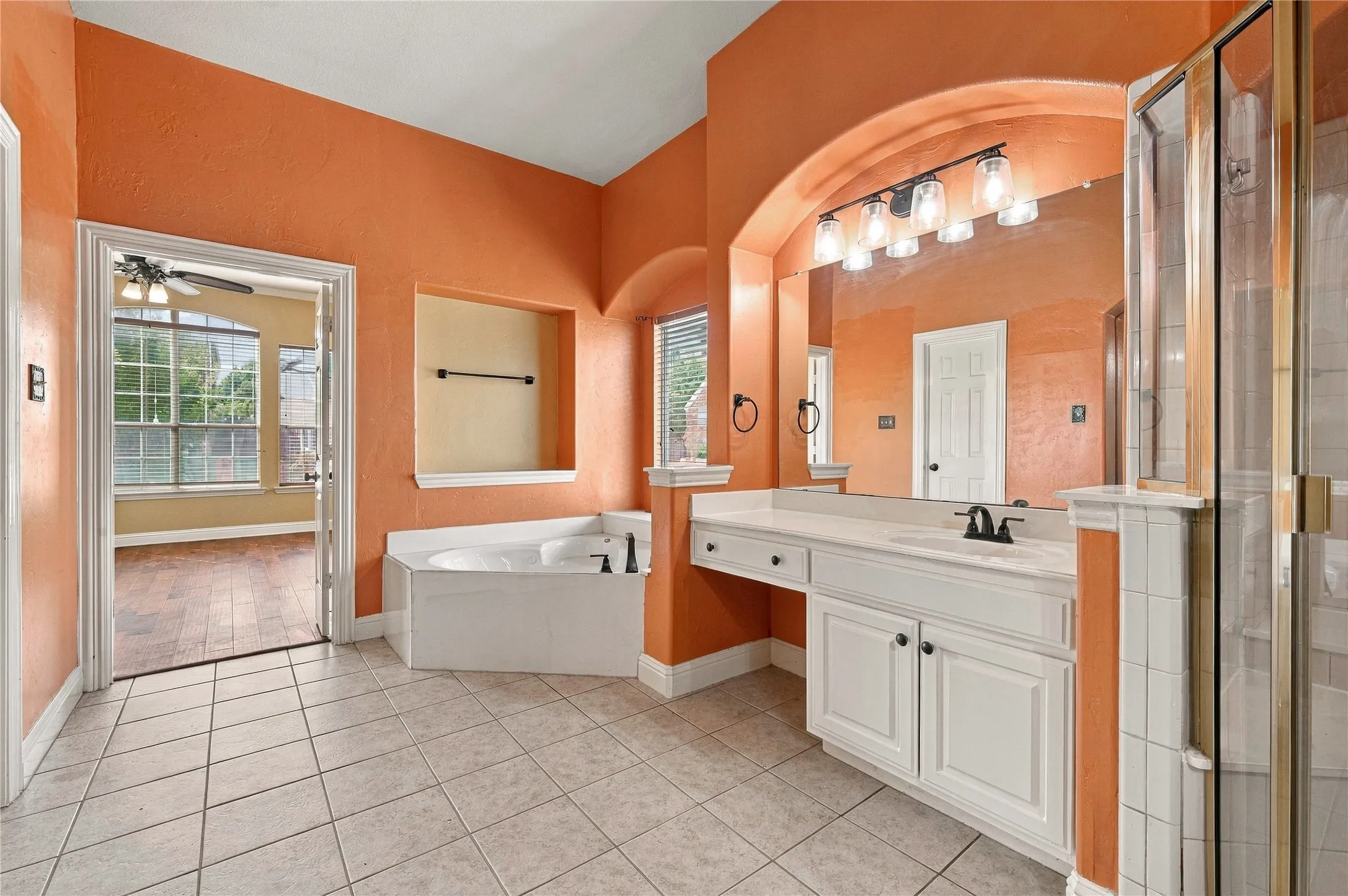 Tiled flooring, built-in corner bathtub, vanity with dual sinks, arched mirror, and separate glass-enclosed shower