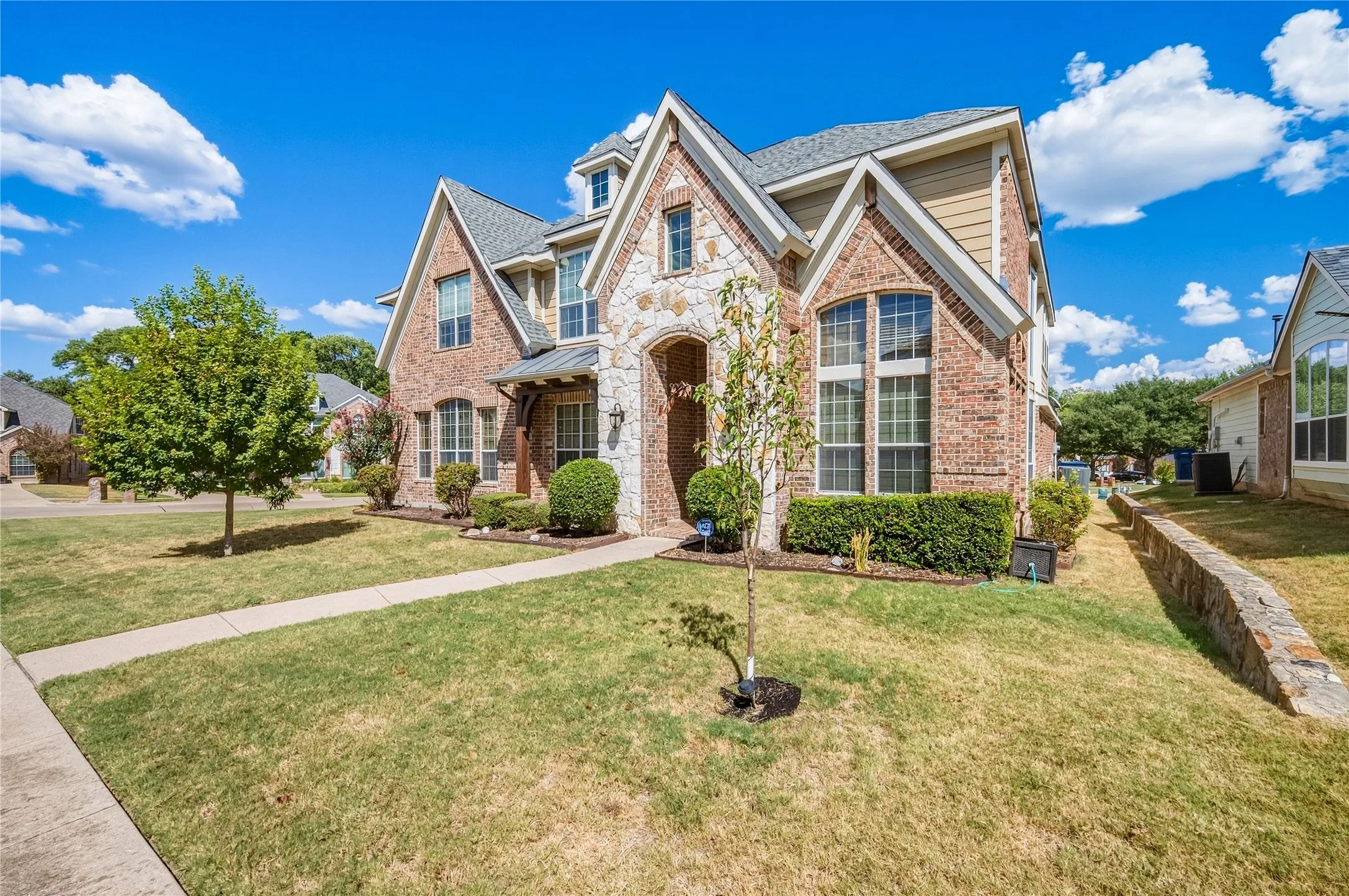 Brick and stone facade, arched entryway, gabled rooflines, decorative window trim, and mature landscaping