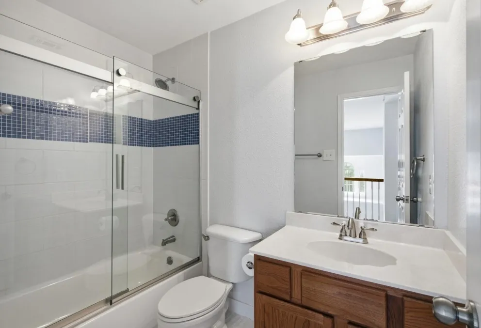 Tub/shower combination with sliding glass doors and blue mosaic tile accent, wood-finish vanity with white countertop, and wall-mounted mirror with overhead light fixture