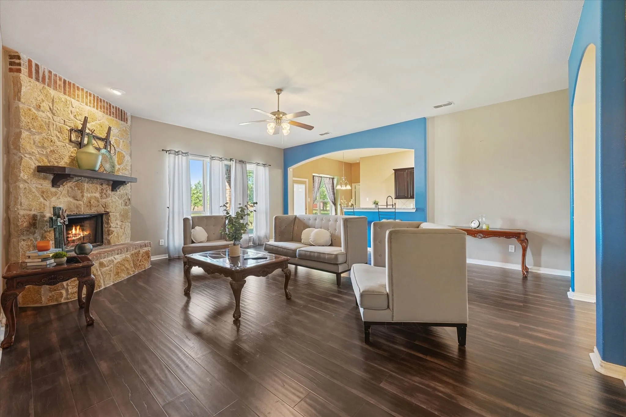 Stone-clad fireplace with wood mantel, dark wood-finish flooring, ceiling fan, and arched wall openings