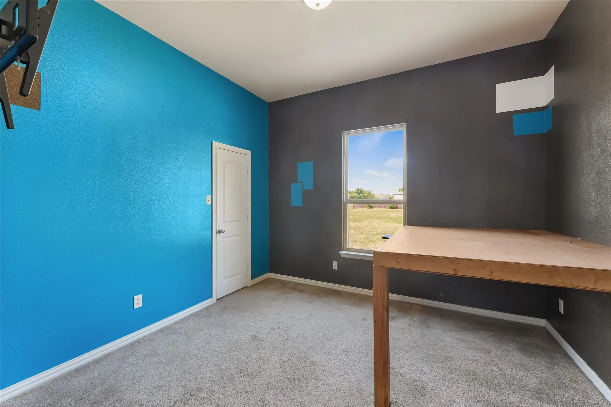 Carpeted room with a single window, a light wood-finish built-in desk, and a door with white trim