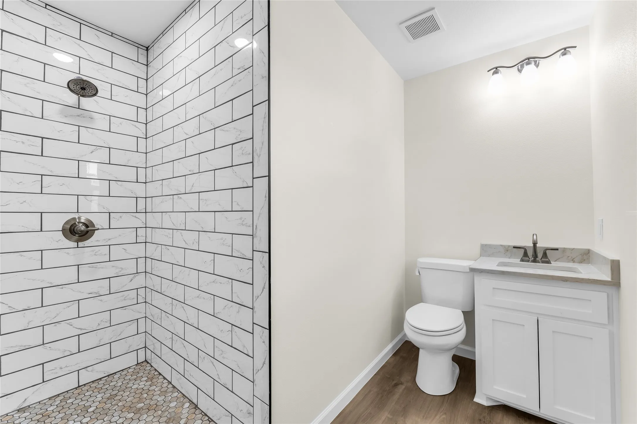 White subway tile shower with a mosaic tile floor, wood-finish flooring, white vanity with stone countertop, and contemporary track lighting