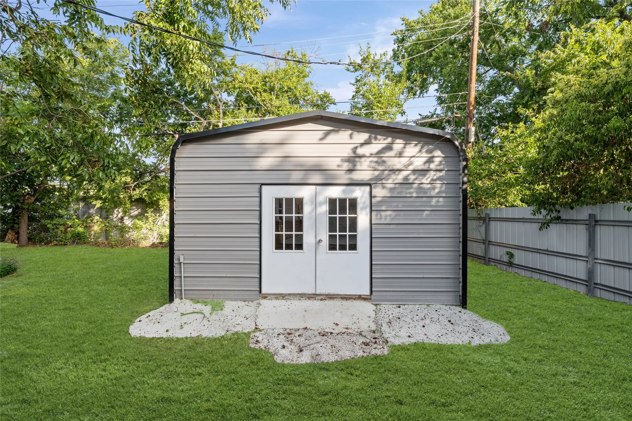 Corrugated metal storage shed with double doors featuring grid-patterned glass panels, set on a concrete pad, surrounded by a manicured lawn
