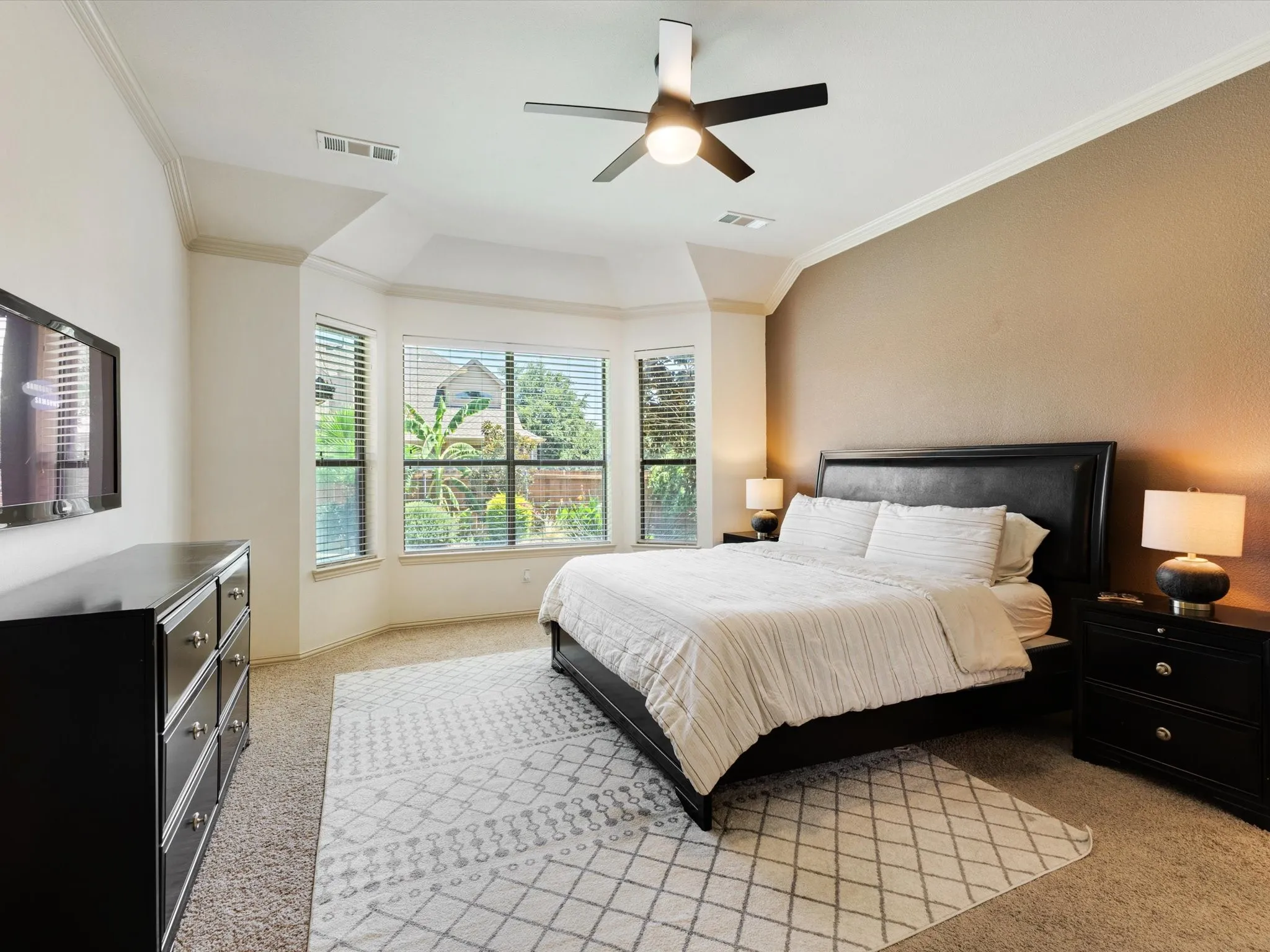 Tray ceiling, ceiling fan with integrated lighting, bay window, textured accent wall, and crown molding