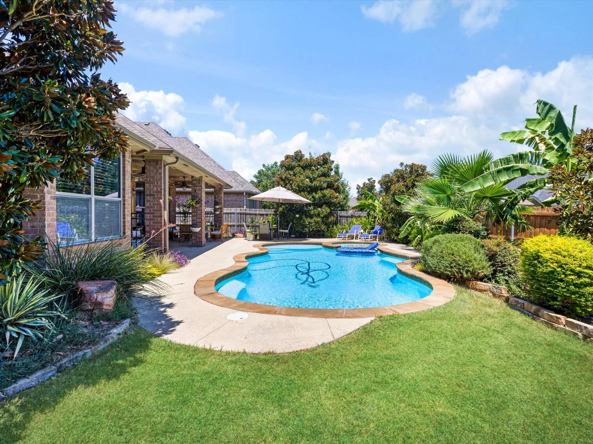 Curved swimming pool with an integrated spa, expansive brick patio, lush landscaping, and a covered outdoor living area