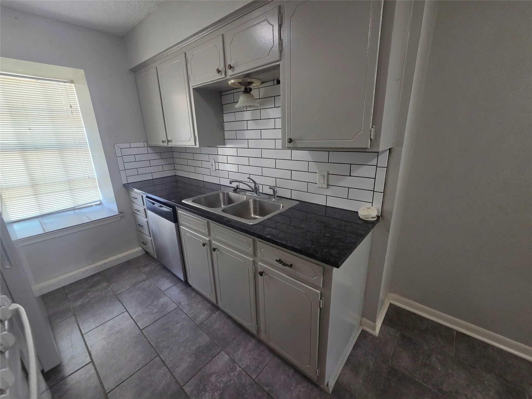 Kitchen featuring white cabinetry, dark countertops, a subway tile backsplash, a stainless steel dishwasher, and tile flooring